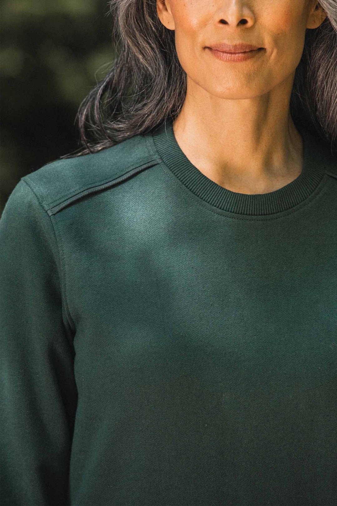 A woman with long, gray hair and a slight smile stands outdoors wearing the Bridge & Burn Bailey Crew Sweatshirt in Juniper.