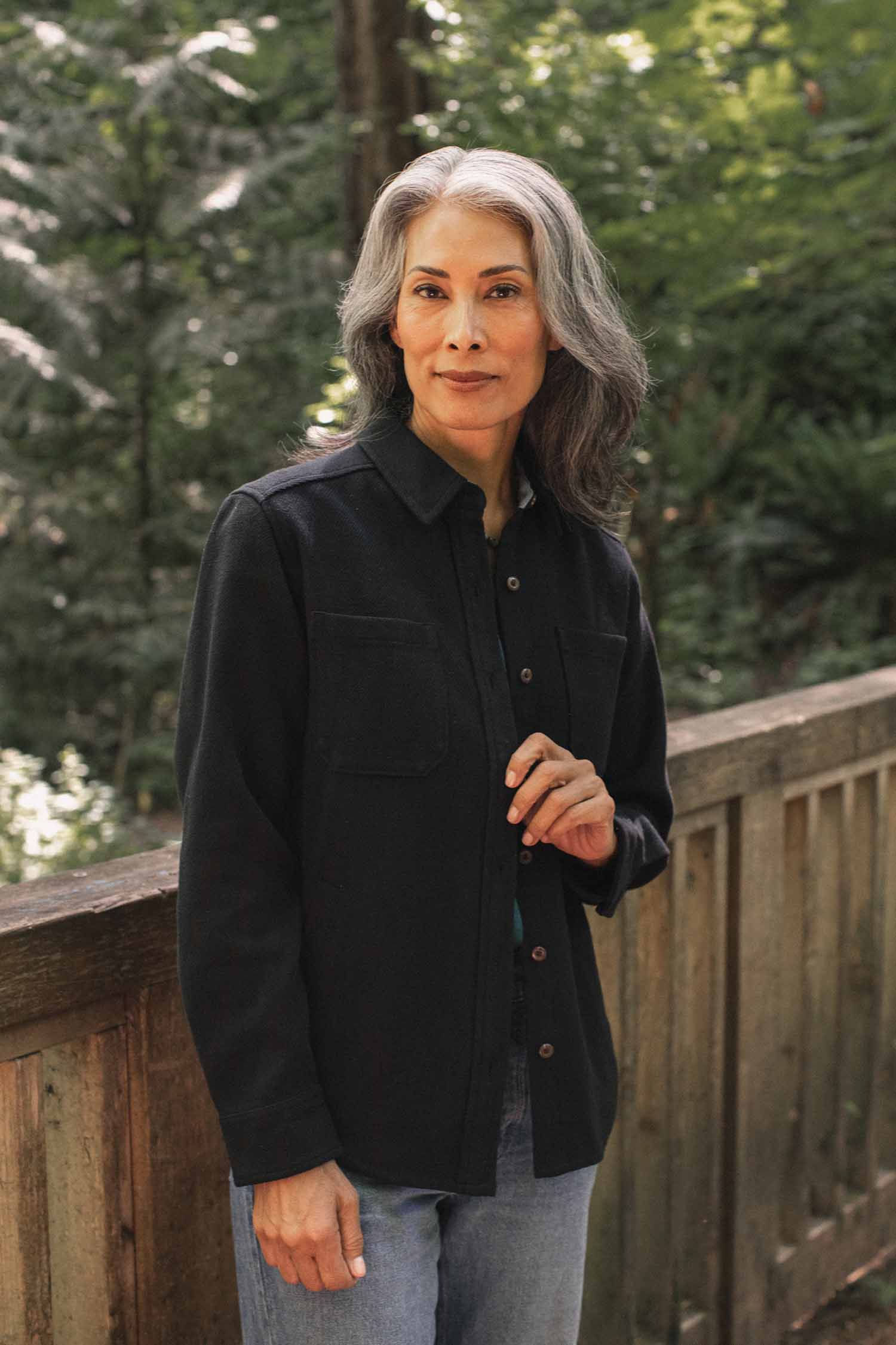A woman with long gray hair, dressed in the Bridge & Burn Dawson Overshirt in Ink Stone and jeans, stands outdoors on a wooden bridge with a forest backdrop.