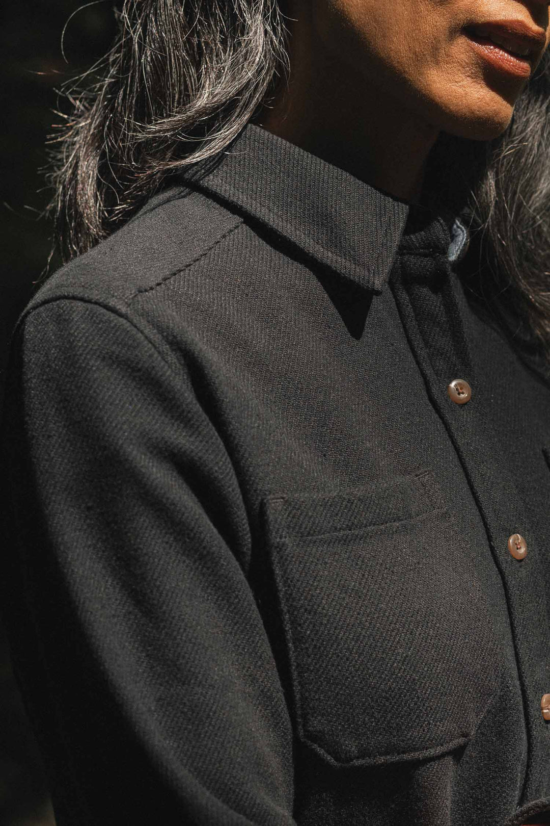 A person with long, dark hair stands in sunlight, wearing the Bridge & Burn Dawson Overshirt in Ink Stone—a black recycled overshirt featuring brown buttons and chest pockets. Their face is partially visible against an out-of-focus background.