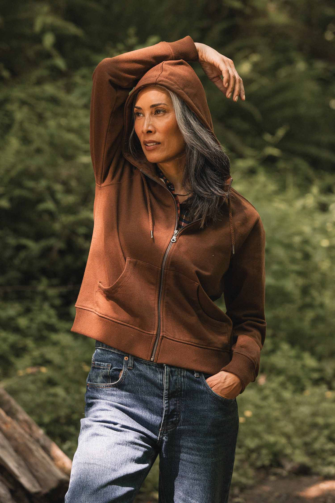 A woman with long, gray-streaked hair wears the Bridge & Burn Mason Hoodie in Cumin and jeans, standing outdoors against a backdrop of greenery, one hand in her pocket and the other raised to touch her hood.