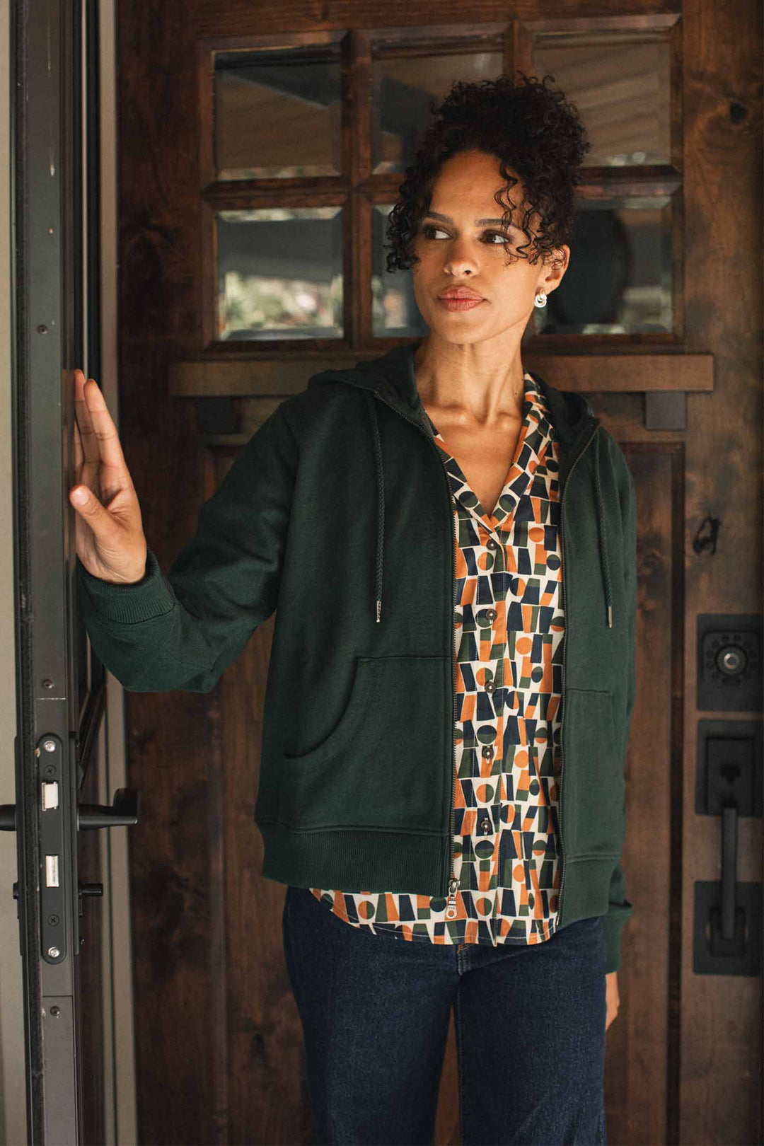 A woman with curly hair stands by a wooden door, wearing the Bridge & Burn Mason Hoodie in Juniper—a dark green sustainable layer crafted from recycled cotton—over a patterned shirt and jeans, looking thoughtfully to the side with one hand resting on the doorframe.