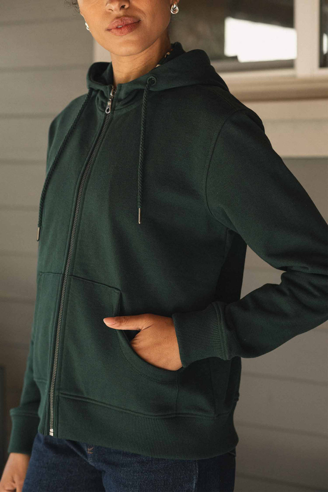 A person wears the Bridge & Burn Mason Hoodie in Juniper—a dark green sustainable piece crafted from recycled cotton—featuring a drawstring hood, visible zipper, and one hand casually placed in the pocket while standing before a light grey wall.
