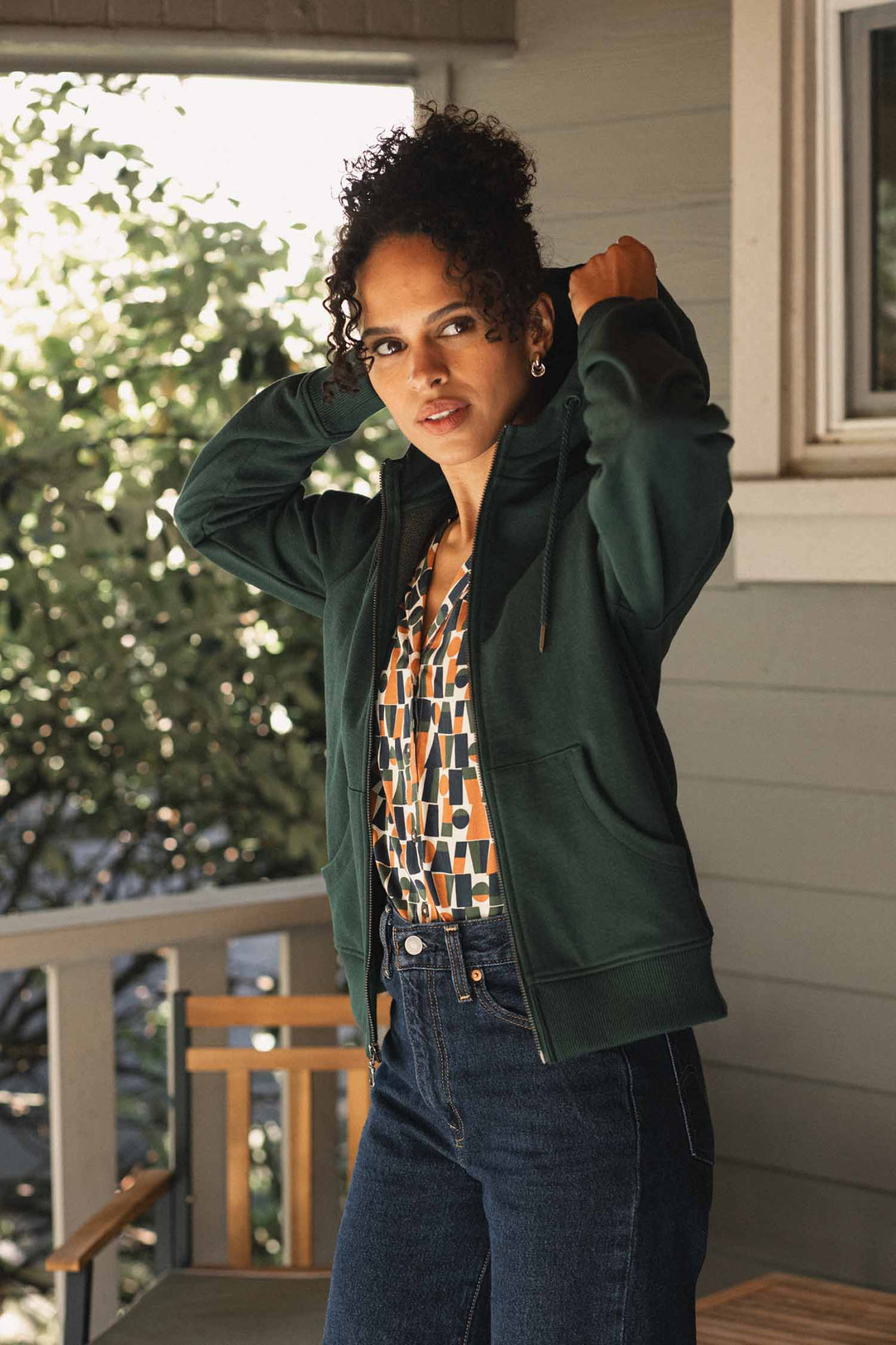 Wearing the Mason Hoodie in Juniper by Bridge & Burn, a person stands on a porch adjusting their hood and looking at the camera. Made from recycled cotton, the sustainable green hoodie is complemented by trees and part of a window in the background.