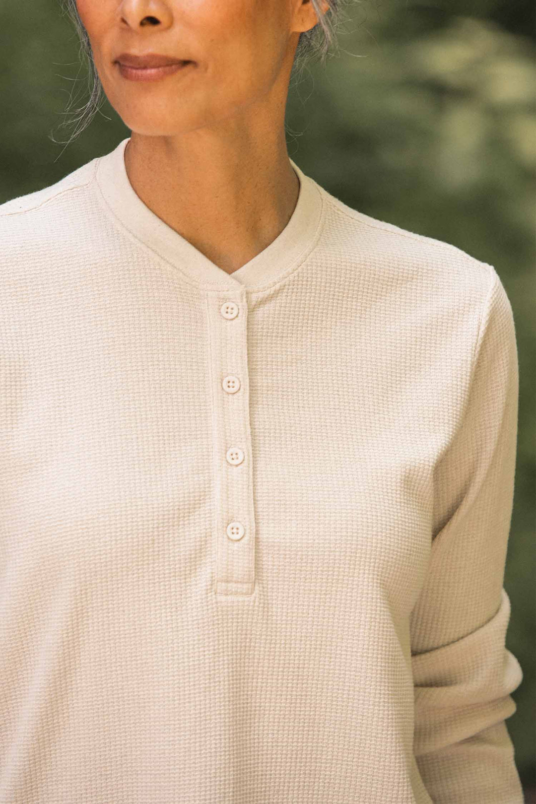 A woman stands outdoors, partially smiling, wearing the Bridge & Burn Women’s Recycled Waffle Henley in Ivory—a long-sleeved shirt crafted from recycled fibers with classic button details.
