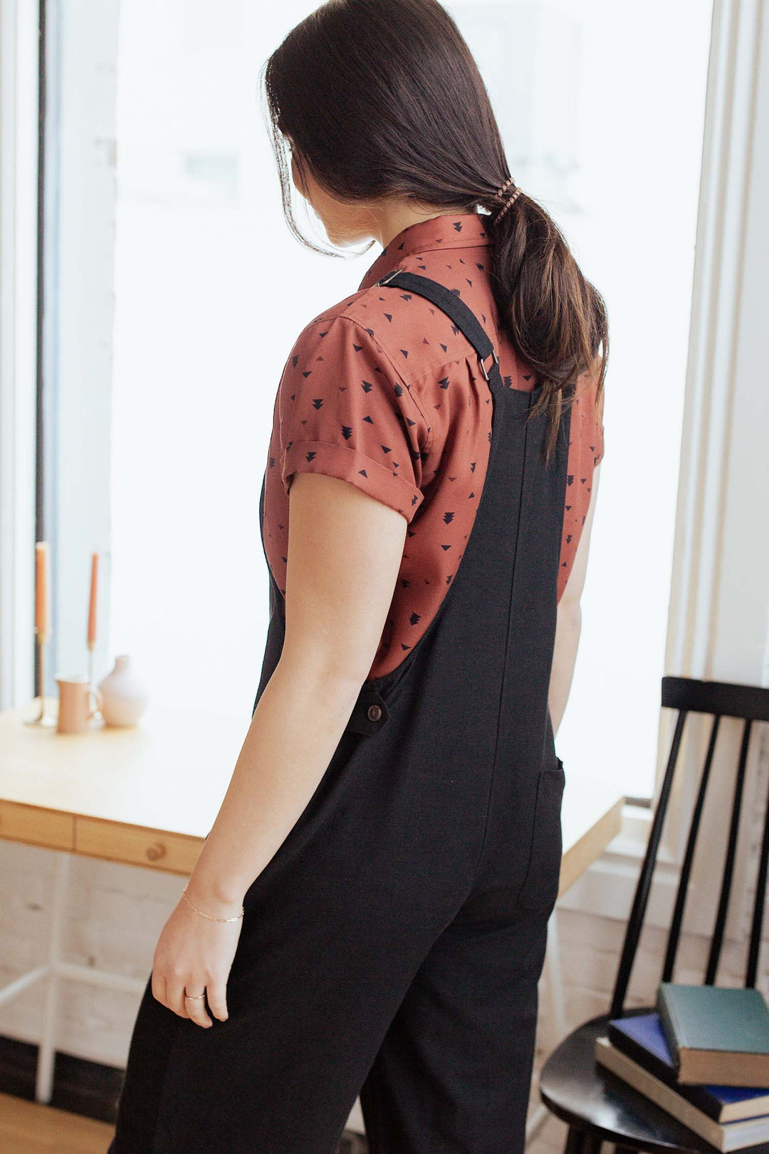 A woman with dark hair in a ponytail stands indoors, facing away. She wears a rust-colored shirt with black hearts, styled with the Bridge & Burn Edie Overall in Black—a relaxed fit linen jumpsuit featuring adjustable straps. A wood table, candles, and a black chair are visible nearby.