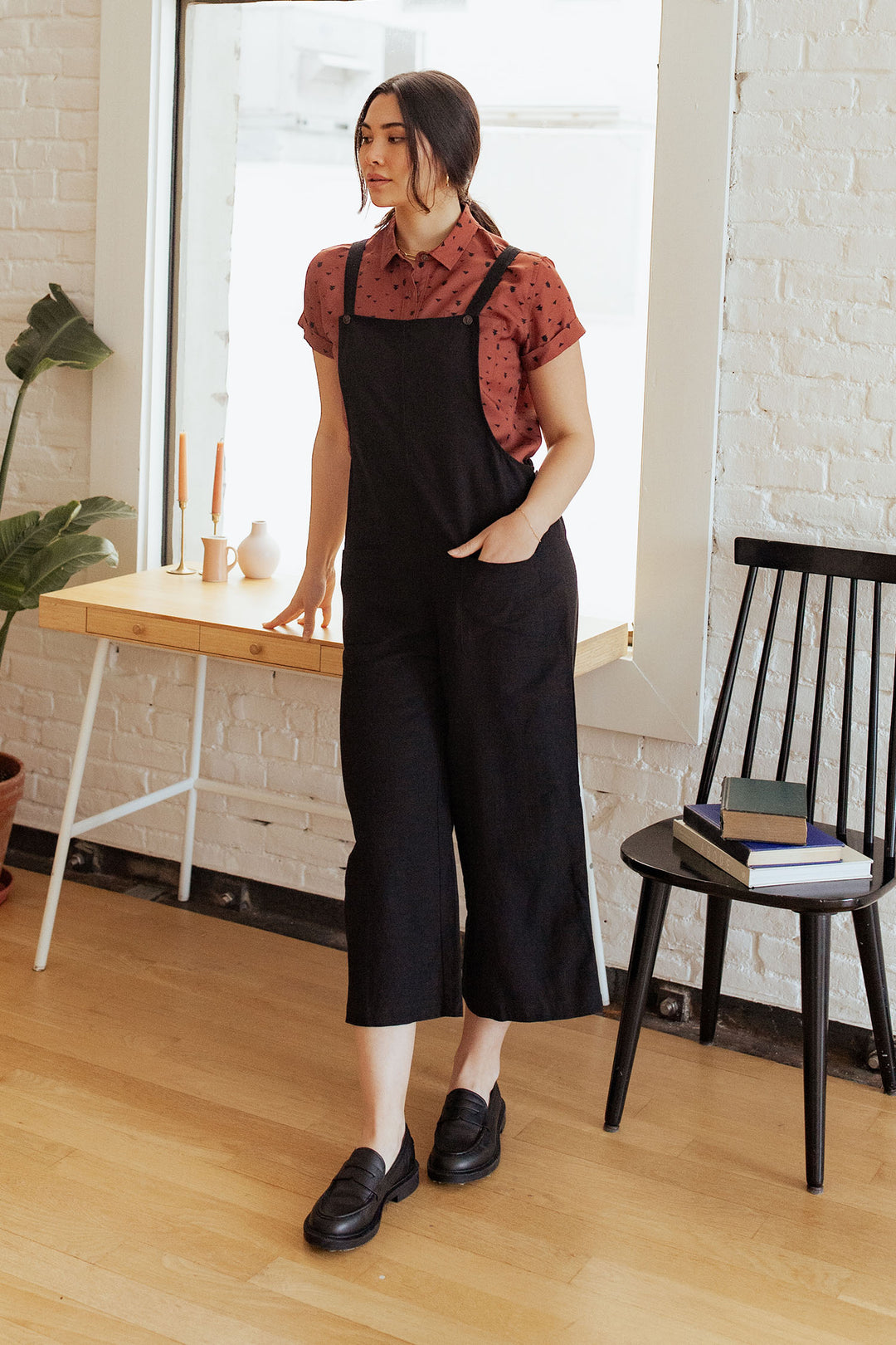 A woman stands indoors by a desk, wearing the Bridge & Burn Edie Overall in black—a relaxed fit linen jumpsuit with adjustable straps—layered over a red patterned short-sleeve shirt and paired with black shoes. Nearby, a black chair holds books, and a small table displays candles and a vase.