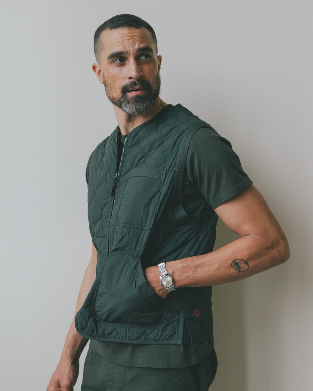 A man wearing an Imperfects - Shokunin Vest in Quilted Nylon / Greenroom over a matching dark green t-shirt, with his hand in the front pocket.