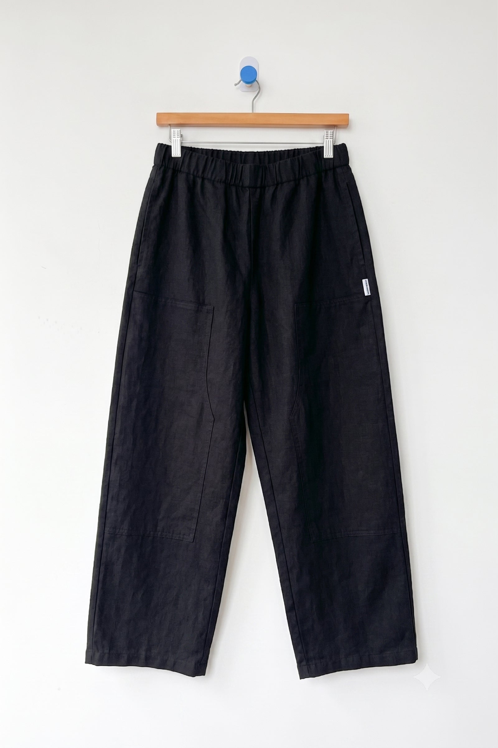 Le Bon Shoppe’s Gardener’s Pant in Black features a wide-leg linen silhouette with an elastic waistband and spacious front pockets, designed for a relaxed fit. Shown here hanging on a wooden hanger against a plain white wall.
