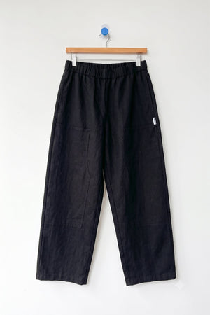 Le Bon Shoppe’s Gardener’s Pant in Black features a wide-leg linen silhouette with an elastic waistband and spacious front pockets, designed for a relaxed fit. Shown here hanging on a wooden hanger against a plain white wall.