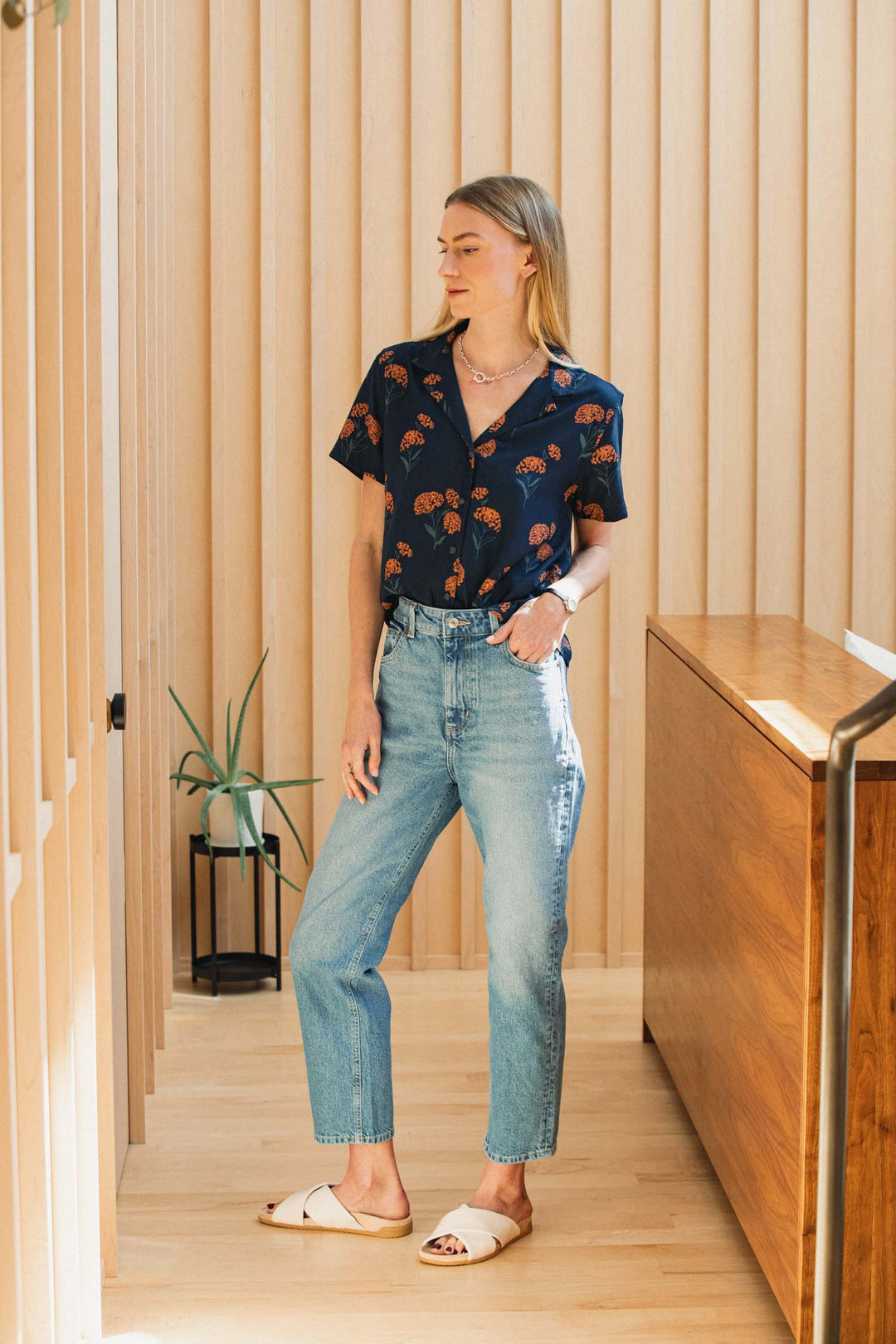 A woman wears a navy blue Bridge & Burn Mila Cropped Shirt / Wildflower tucked into high-waisted blue jeans and cream-colored slide sandals.