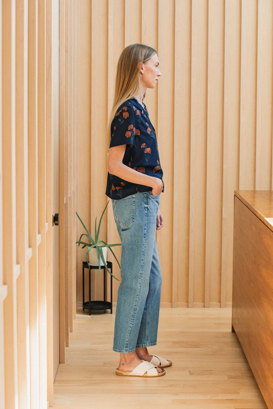 A person wearing a navy Bridge & Burn Mila Cropped Shirt / Wildflower with a rust-colored wildflower pattern, light-wash jeans, and white slide sandals.