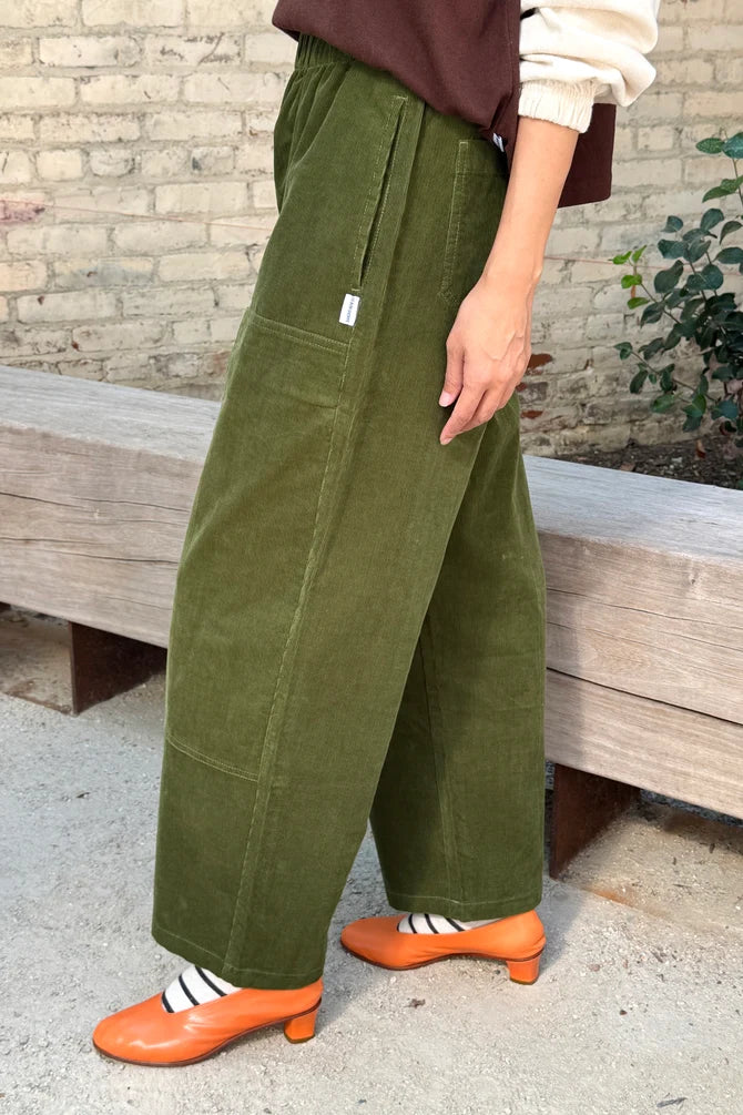 Side view of Le Bon Shoppe Gardener’s Pant in Bay Leaf corduroy, featuring wide-leg styling and large patch pockets, worn with orange block-heeled shoes and striped socks.