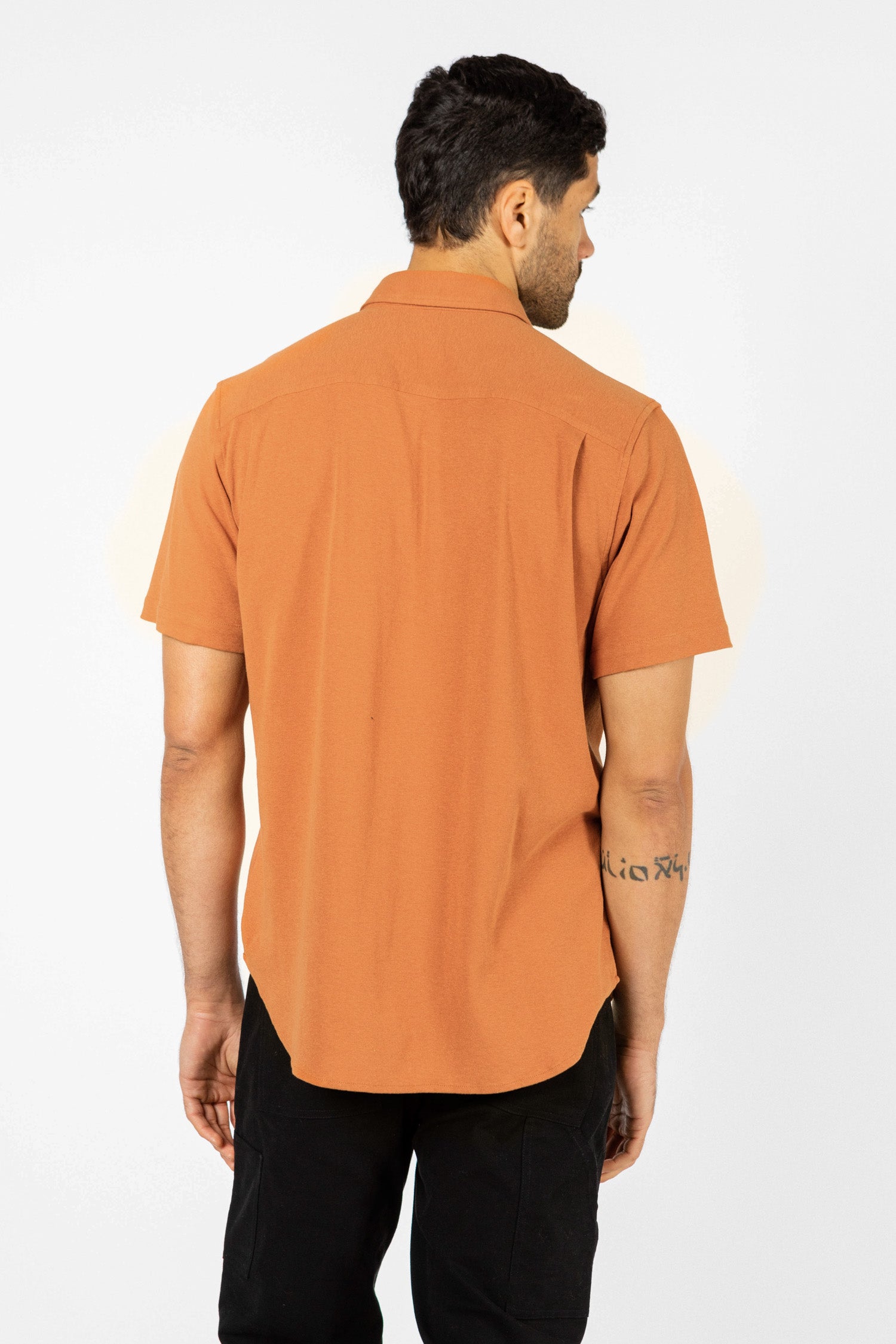 A man stands with his back to the camera, modeling Bridge & Burn’s Gary Knit Pullover in Clay Red Jersey and black pants against a plain white background.