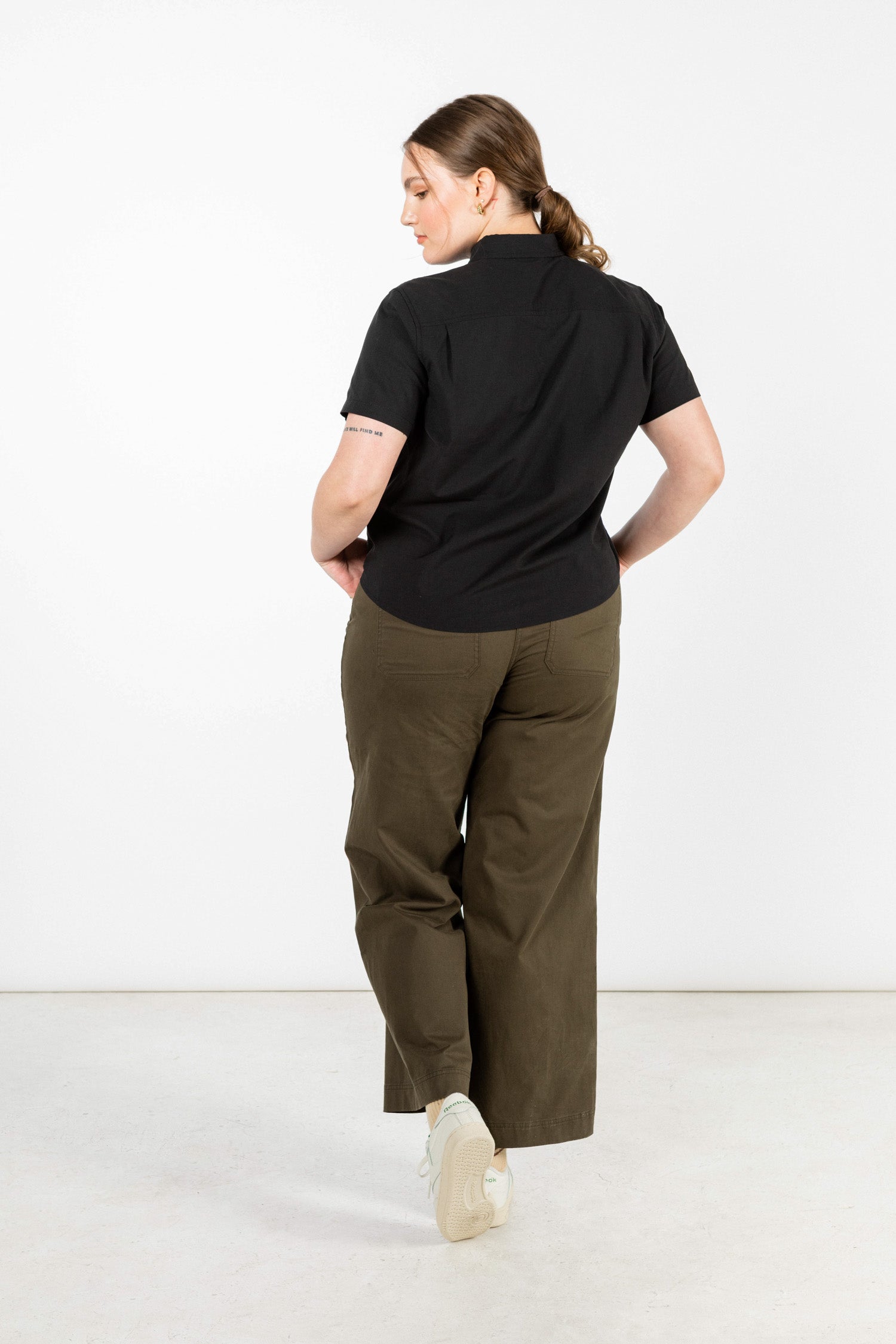 A person with light skin and brown hair in a low ponytail stands facing away, wearing the Bridge & Burn Greer Cropped Shirt in Black Poplin, olive green wide-leg pants, and white sneakers against a plain white background.