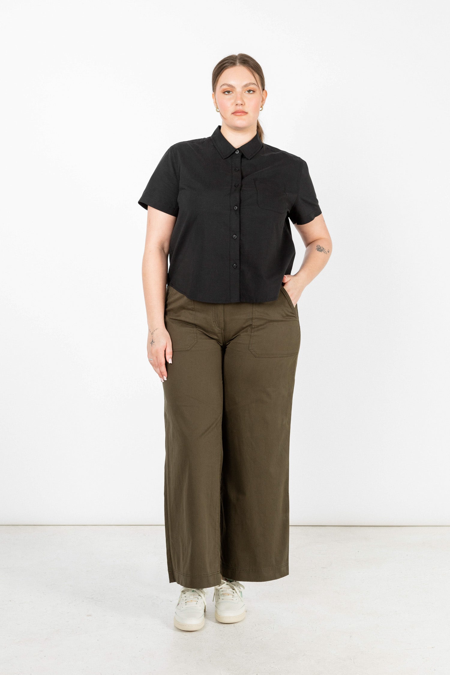Against a white background, a person with light brown hair pulled back wears Bridge & Burn's Greer Cropped Shirt in black poplin, olive green wide-leg pants, and white sneakers—one hand is in their pocket.