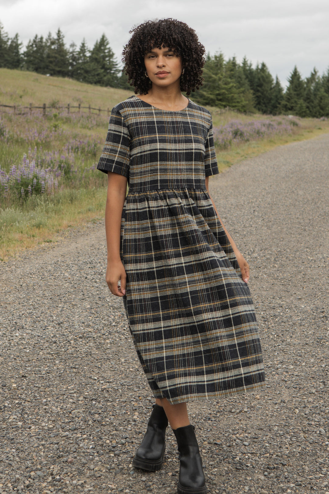 Wearing the Perry Dress / Volcanic Plaid by Bridge & Burn, a person with curly hair stands on a gravel road lined with wildflowers and trees under a cloudy sky, pairing the midi-length recycled cotton dress with black boots.