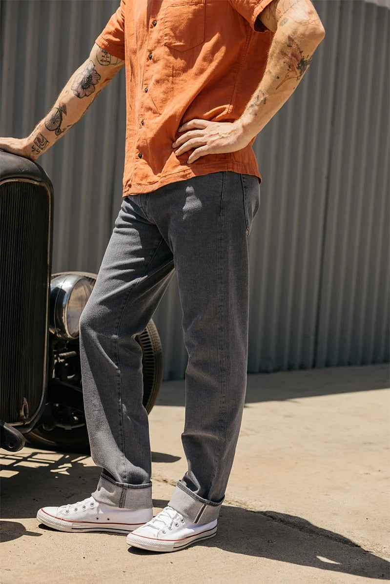 A person with tattoos, wearing an orange short-sleeve shirt, Hiroshi Kato - The Hammer Straight / Granite 10.5oz Selvedge jeans cuffed at the ankles, and white Converse sneakers stands outdoors, leaning on an old car. A corrugated metal wall serves as the backdrop.