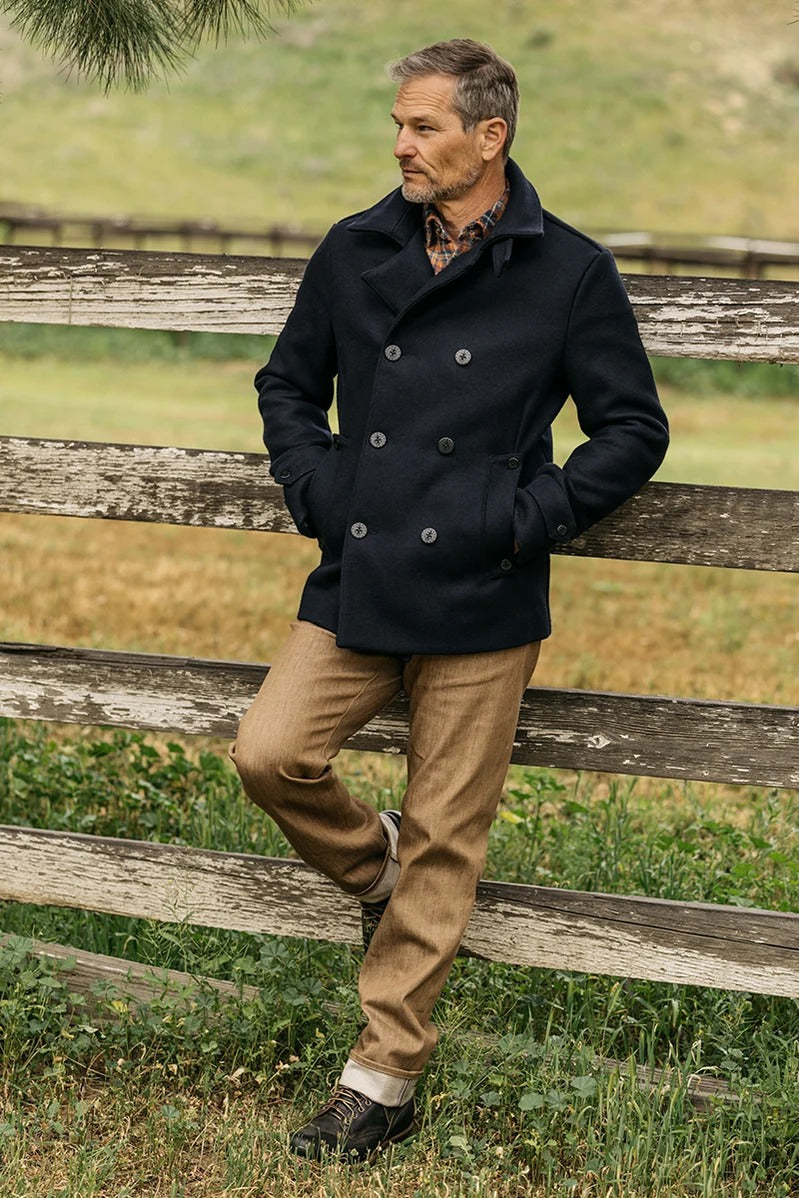 A man in a navy peacoat, brown pants, and boots stands outdoors, leaning against a rustic wooden fence with grassy fields in the background. His look is elevated with Hiroshi Kato’s The Hammer Straight / Copper Raw 10.5oz Selvedge denim, adding a modern edge to his classic style.