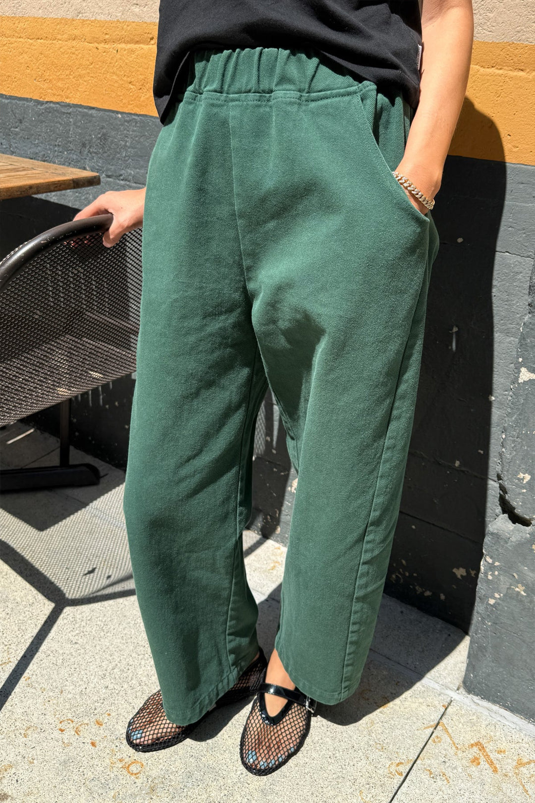 Person stands outdoors in Le Bon Shoppe - Arc Pant / Pine, paired with a black top and black mesh shoes. One hand is in their pocket while the other rests on a metal chair.