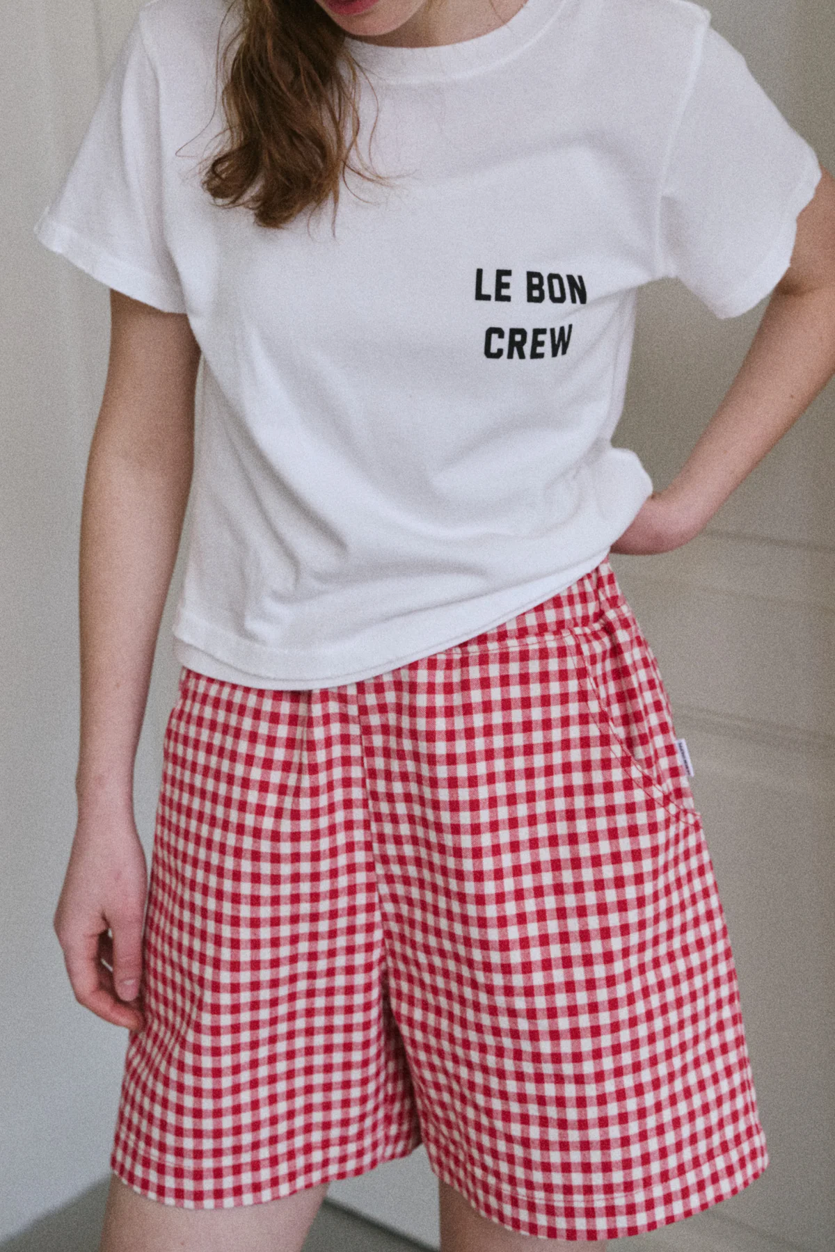A person stands indoors with one hand on their hip, wearing a white LE BON CREW t-shirt and Le Bon Shoppe - City Short in Red Gingham. Their face is partially out of frame.