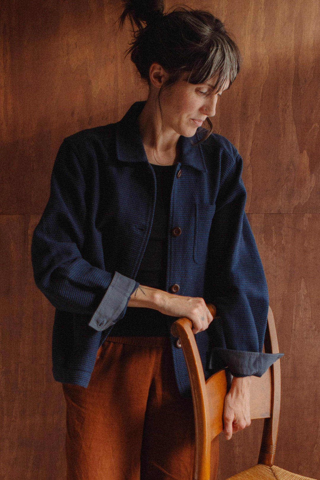 A woman with dark hair and gray streaks stands indoors against a brown background, looking down while wearing the Bridge & Burn Women’s Studio Chore Coat in Indigo—an organic cotton workwear jacket with reinforced elbows—paired with brown pants. She rests one hand on the back of a wooden chair.