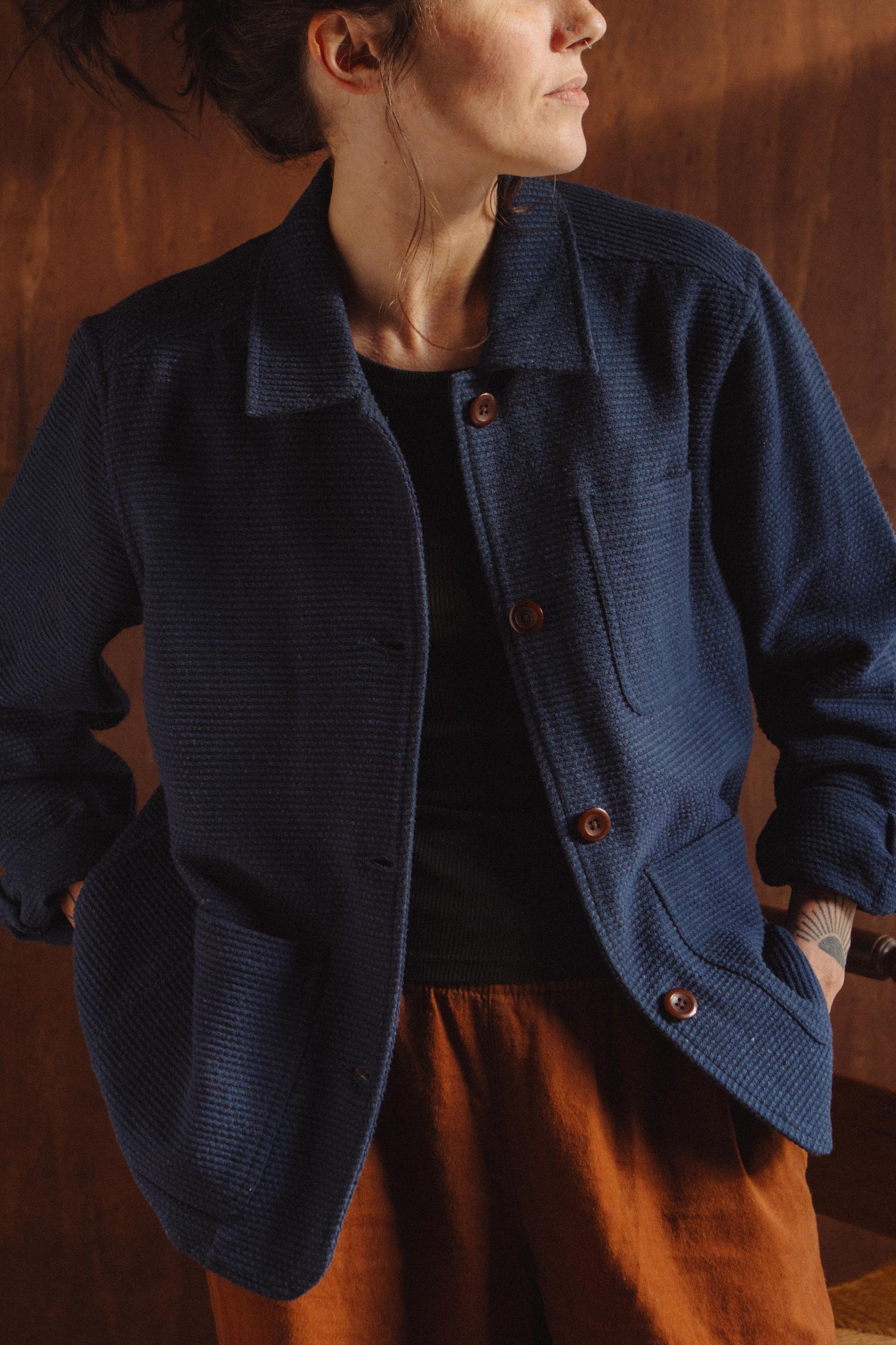 A woman wearing the Bridge & Burn Women’s Studio Chore Coat in indigo—a textured navy blue organic cotton jacket with reinforced elbows—layered over a black shirt and rust-colored pants, stands against a brown background, looking to the side.