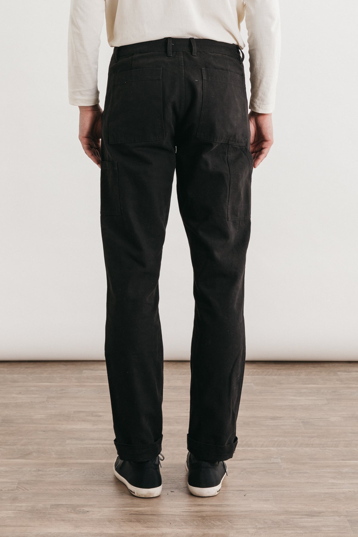 A person stands on a wooden floor, facing away, wearing Bridge & Burn’s Wallace Utility Pant in black canvas, a white long-sleeve shirt, and black shoes against a plain light background.
