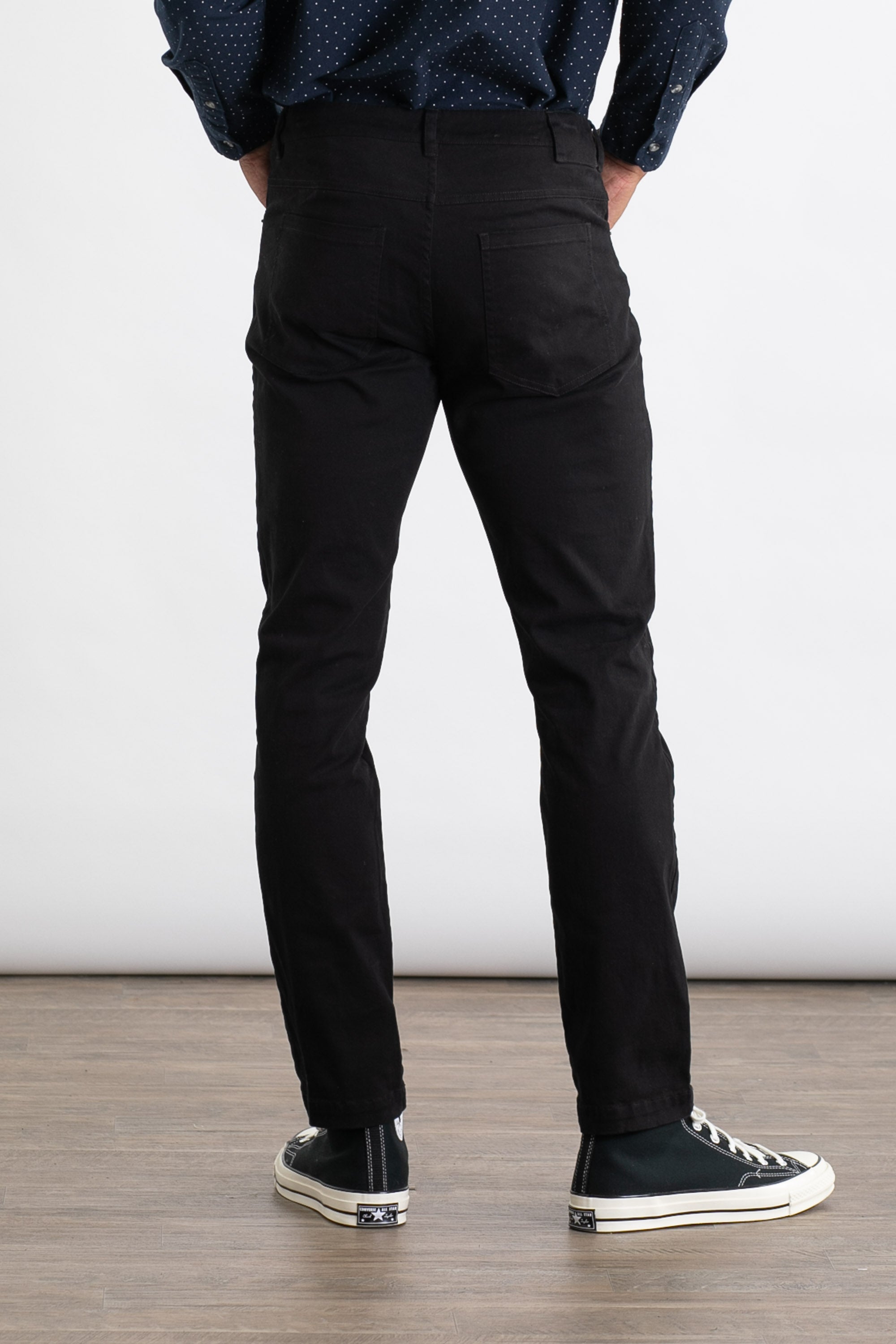 A person stands indoors on a wooden floor, facing away from the camera, wearing Bridge & Burn's Bradley Slim Pant in black with a dark blue long-sleeve shirt with small white dots and black low-top sneakers with white soles.