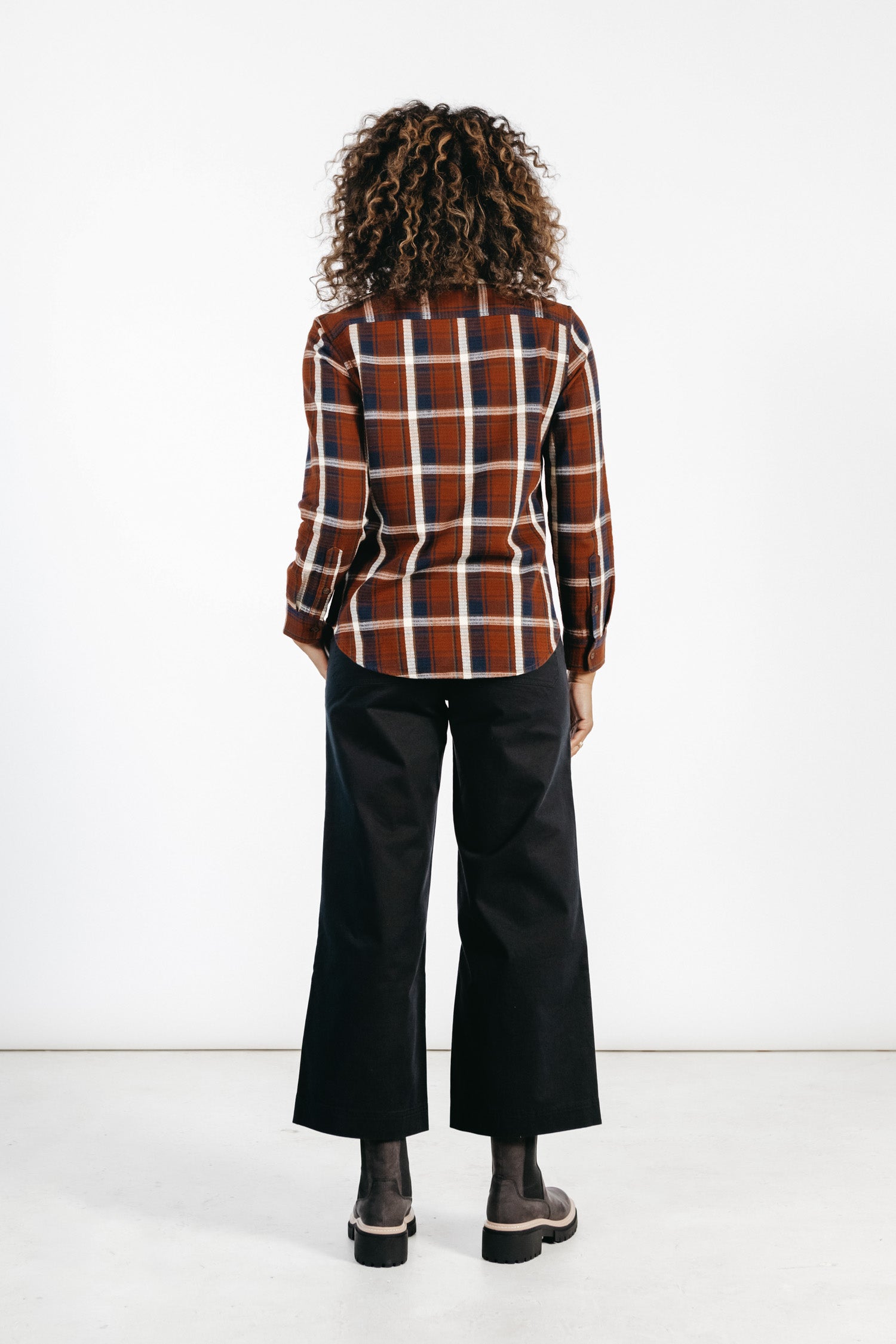 A person with curly hair faces away, wearing the Bridge & Burn Ash Slim Shirt in Echo Ridge Plaid—red and brown—paired with black wide-leg pants and dark shoes, set against a plain white background.