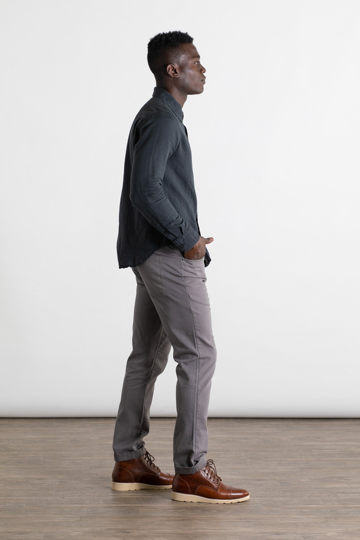 A man stands in profile against a white background, wearing a dark long-sleeve shirt and Bridge & Burn's Bradley Slim Pant in grey—a modern 5-pocket style—paired with brown leather shoes. One hand rests in his pocket as he looks ahead.