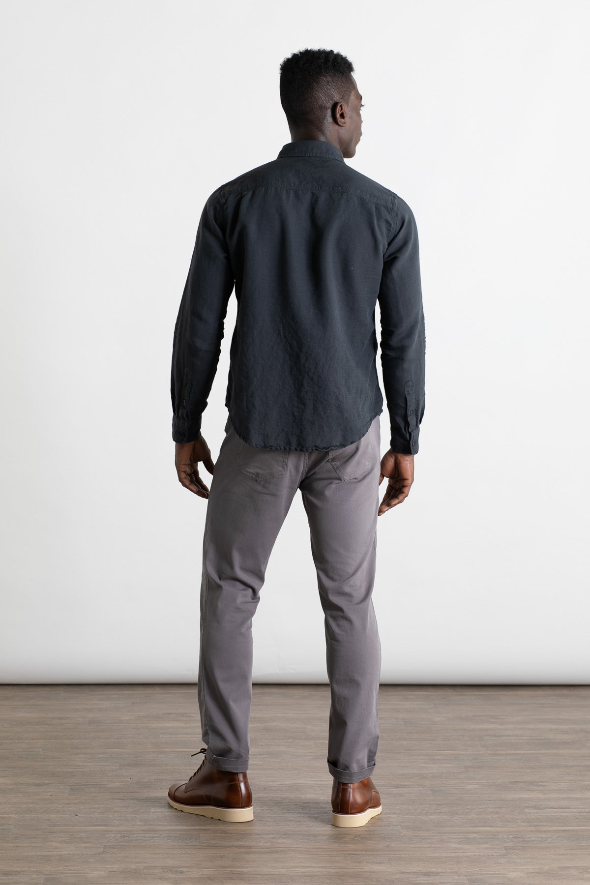 A man stands indoors on a wooden floor with his back to the camera, wearing a dark long-sleeve shirt and Bridge & Burn Bradley Slim Pant in grey. His gray pants and brown shoes stand out against the plain white background.