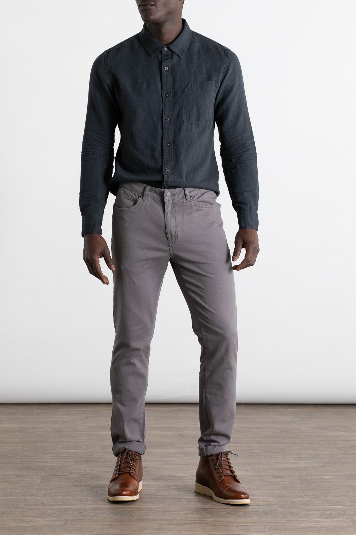 A person stands indoors on a wooden floor wearing Bridge & Burn's Bradley Slim Pant in grey, styled in a 5-pocket design, paired with a dark button-up shirt and brown leather boots; their face is not shown against the plain white background.