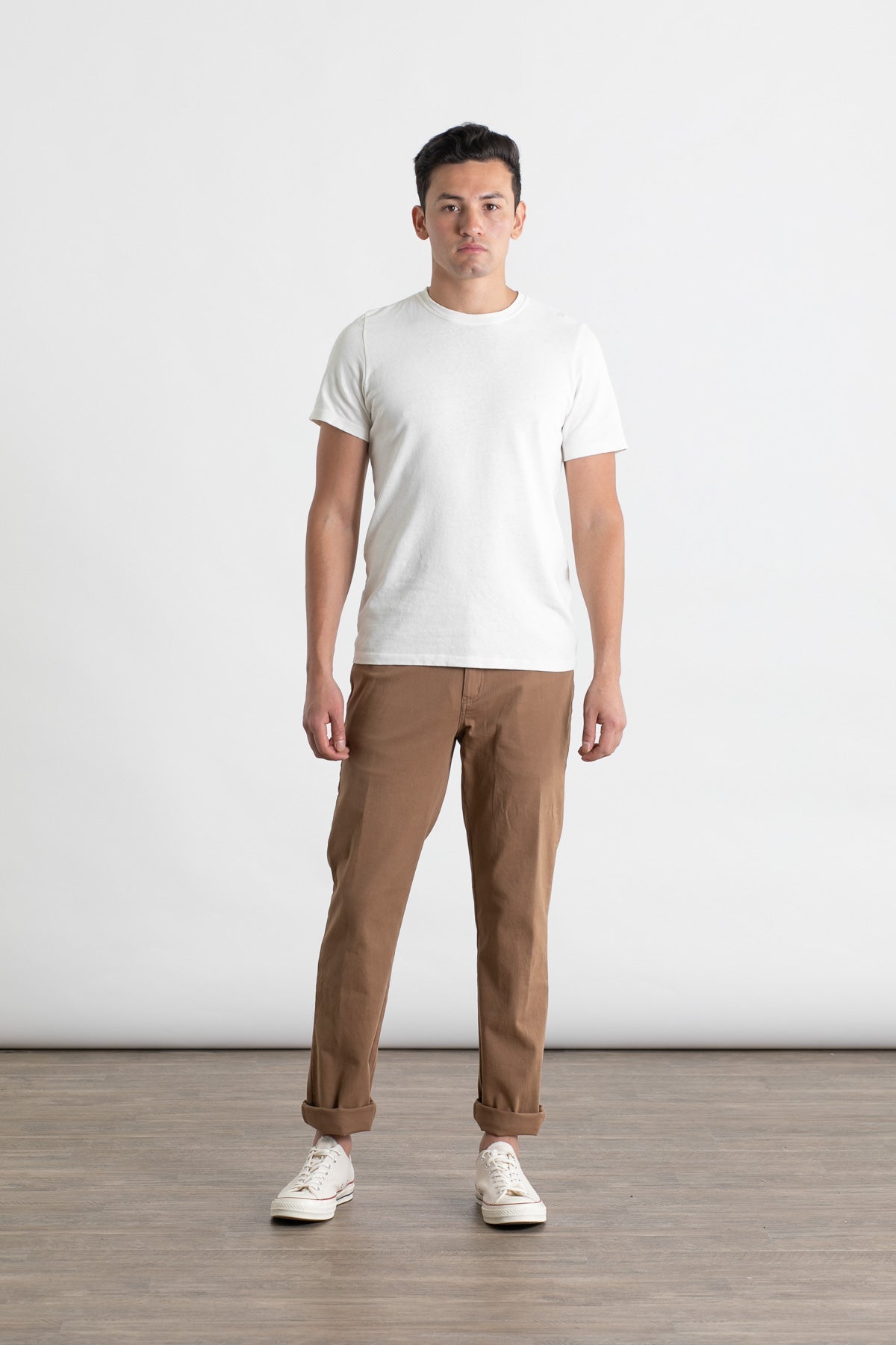 A man stands facing forward against a white background, wearing the Bridge & Burn Polk Pant in brown with 5-pocket styling, a white T-shirt, and white sneakers. He stands on a wooden floor with a neutral expression.