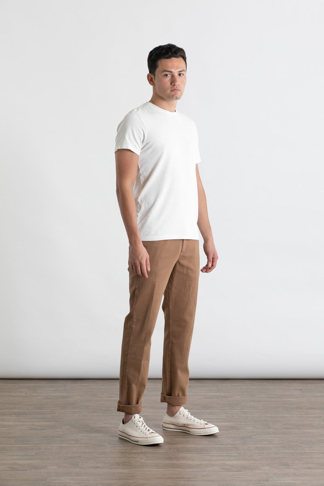 A man stands indoors on a wooden floor against a plain white background, wearing Bridge & Burn's Polk Pant in brown and a white t-shirt. He faces slightly to the side with a neutral expression and wears white sneakers.
