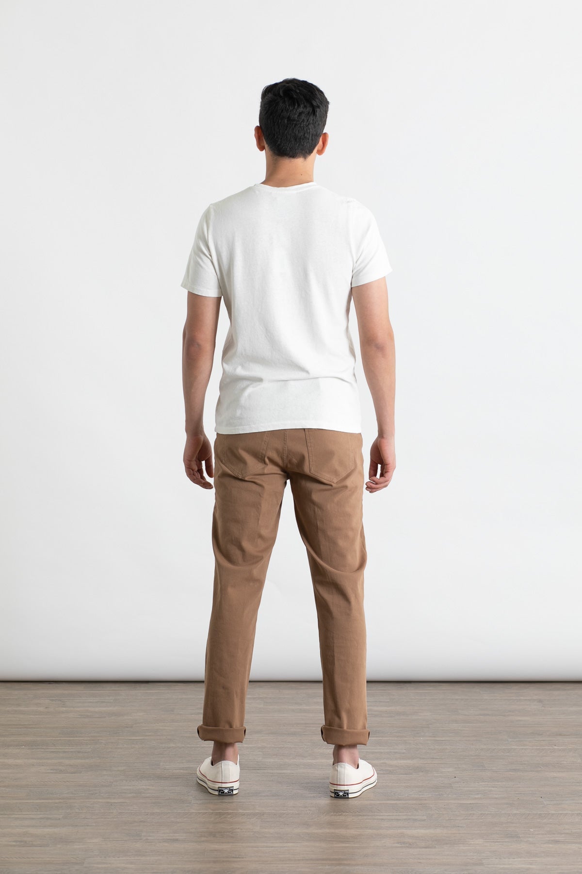A person stands indoors on a wooden floor, facing away from the camera, wearing a white t-shirt and Bridge & Burn’s Polk Pant in brown—straight-fit organic cotton pants with 5-pocket styling. The background is plain.