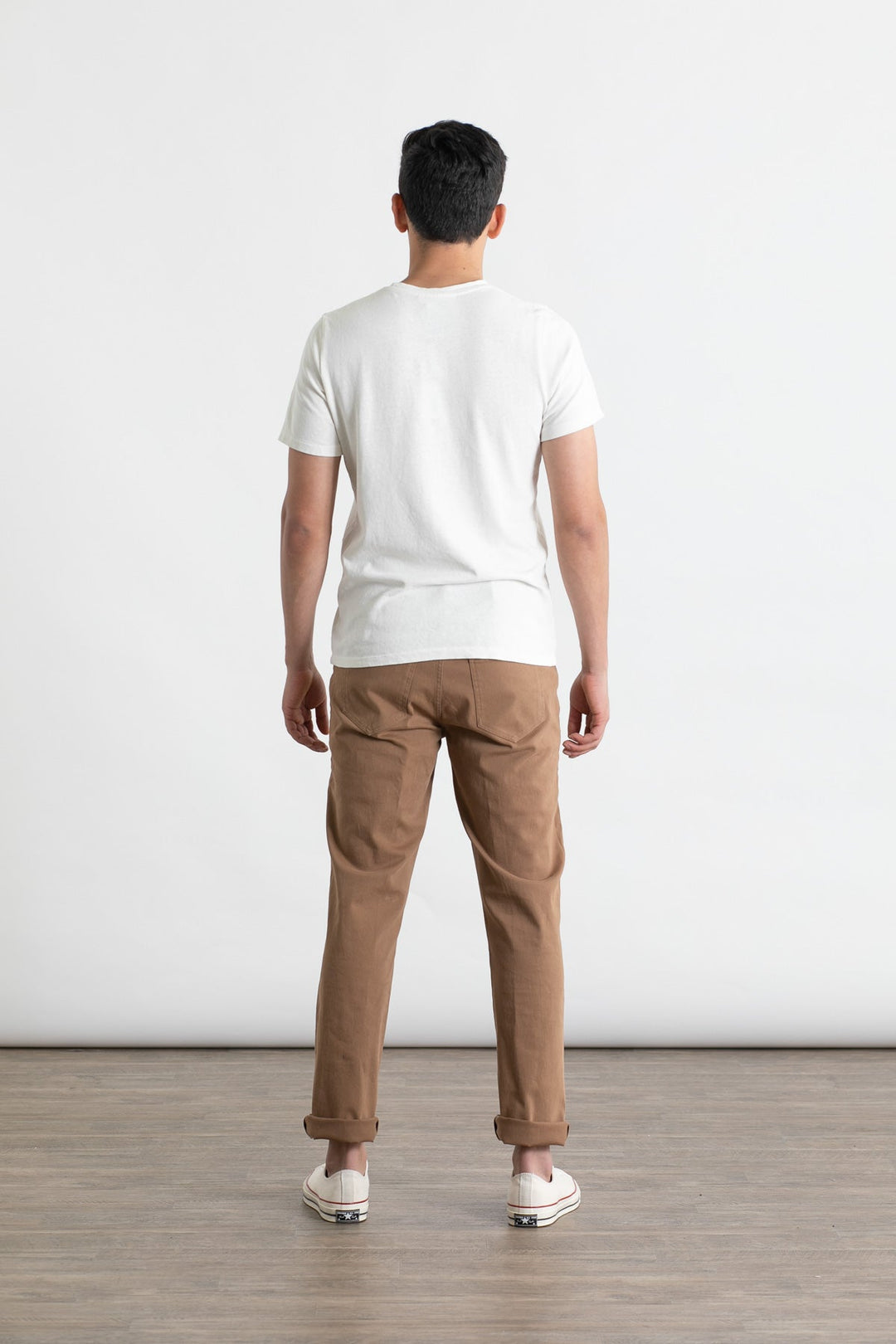 A person stands indoors on a wooden floor, facing away from the camera, wearing a white t-shirt and Bridge & Burn’s Polk Pant in brown—straight-fit organic cotton pants with 5-pocket styling. The background is plain.