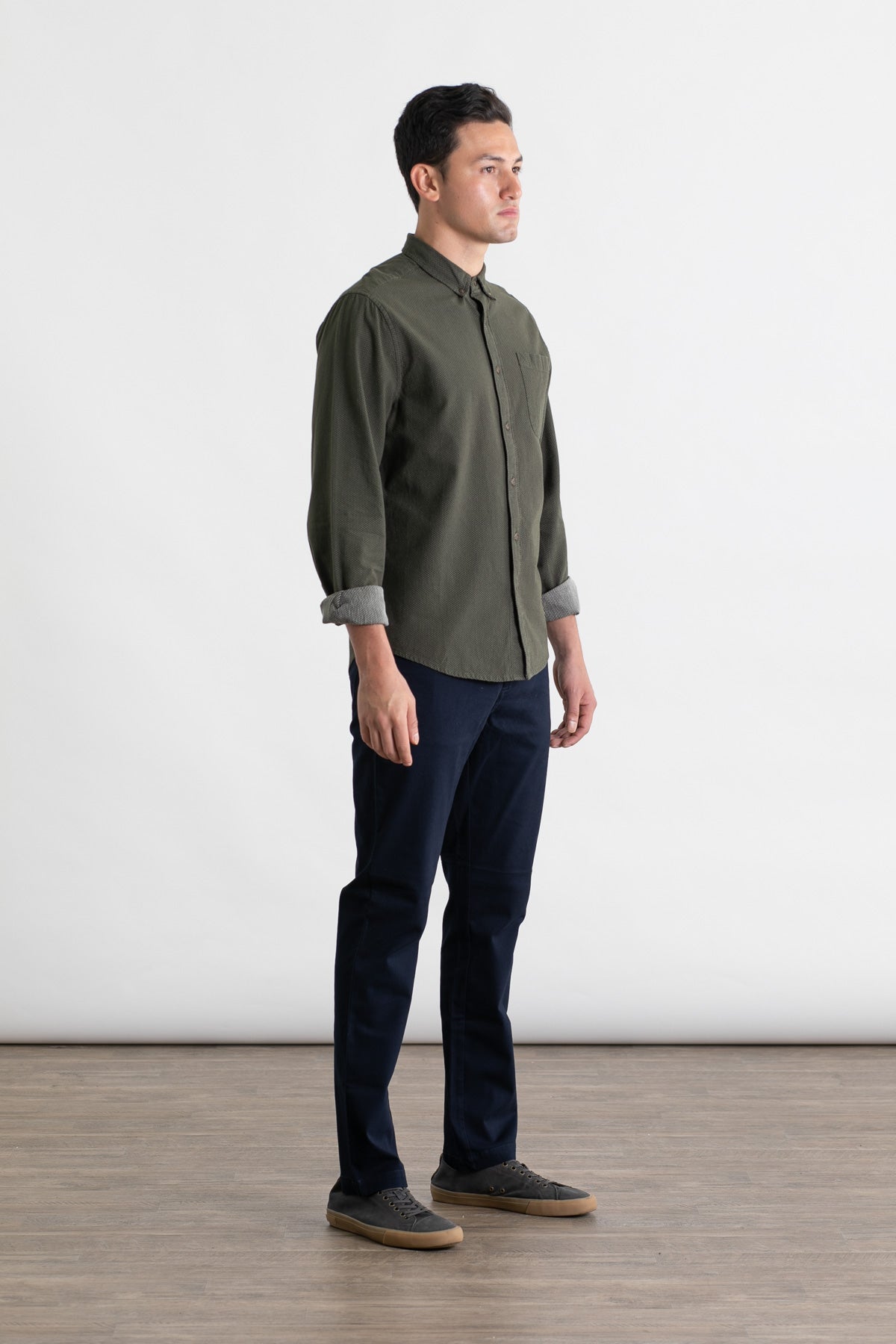 A man stands indoors on a wooden floor against a plain white background, wearing the Bridge & Burn Polk Pant in Navy, a dark green button-up shirt, and gray sneakers. He faces slightly right with a neutral expression.