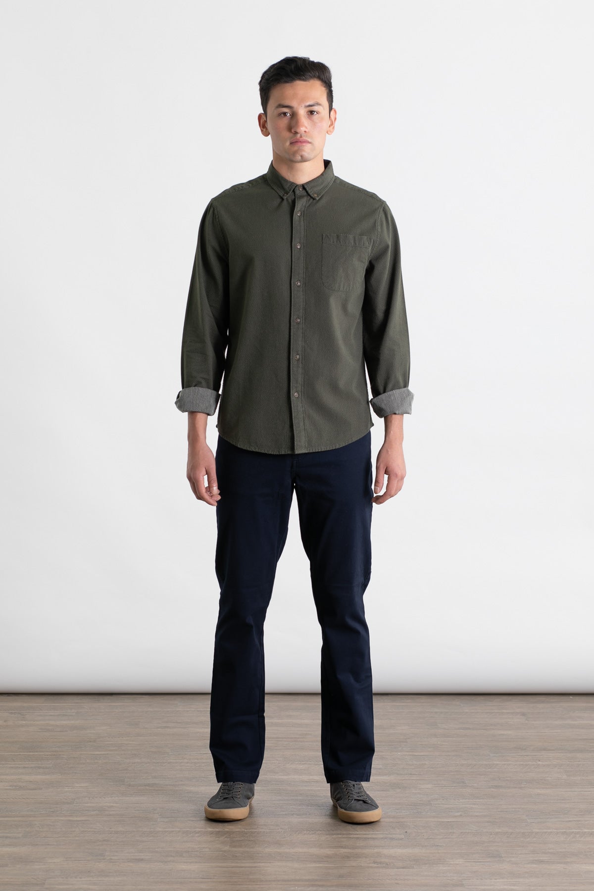 A man stands indoors on a wooden floor against a white background, wearing a dark green button-up shirt, gray sneakers, and Bridge & Burn’s Polk Pant in navy. His arms are relaxed at his sides as he faces forward.