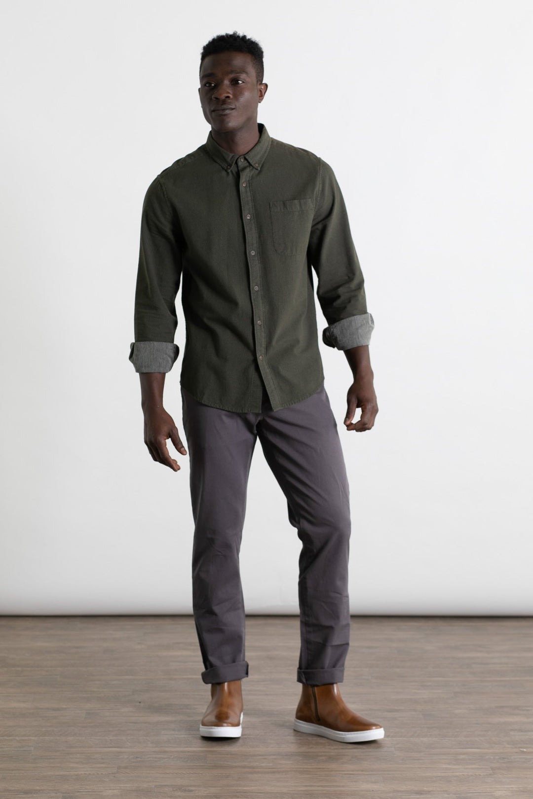 A man stands indoors on a wood floor against a plain white background, wearing a dark green button-up shirt with rolled sleeves, Bridge & Burn Polk Pant in Charcoal, and brown slip-on shoes.