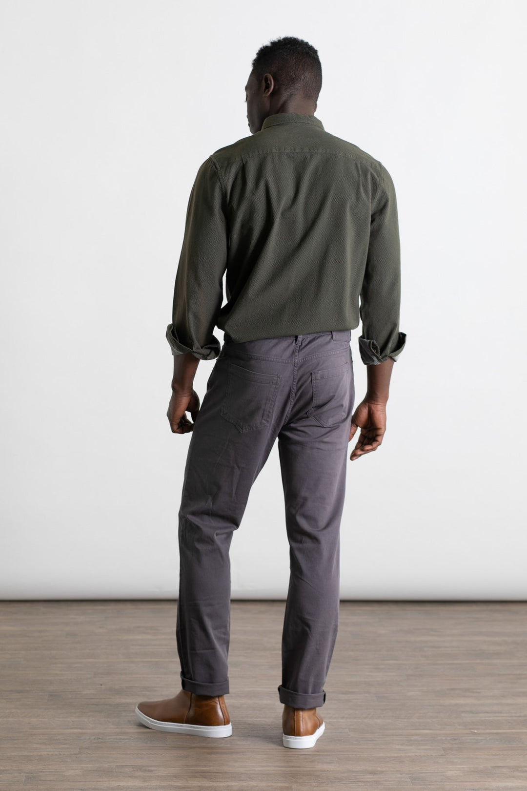 A man stands indoors on a wooden floor, facing away from the camera. He is wearing an olive green long-sleeve shirt, Bridge & Burn Polk Pant in Charcoal, and brown slip-on shoes with white soles. The background is plain and white.