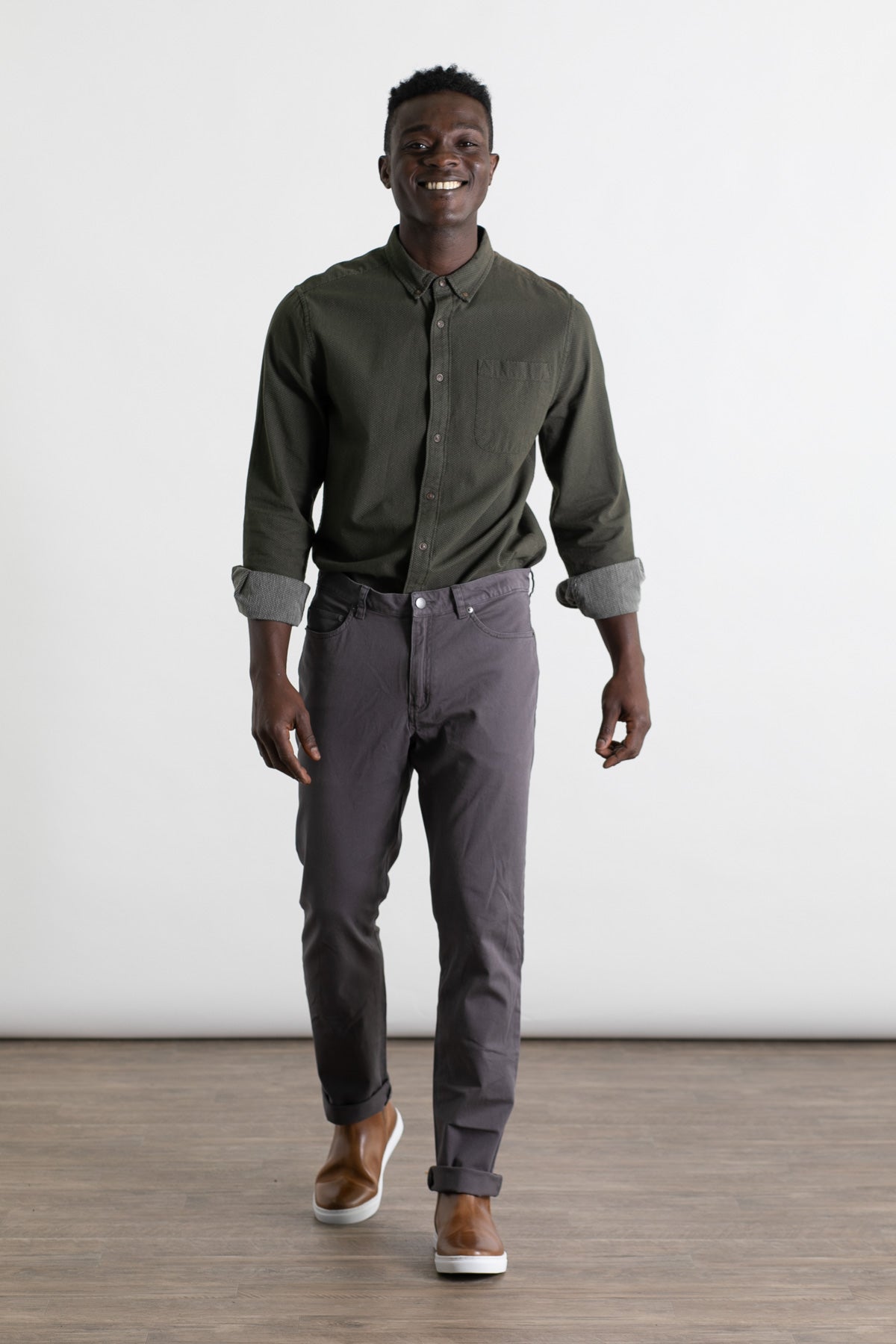 A man with short hair smiles while walking in a well-lit room, wearing the Bridge & Burn Polk Pant in Charcoal, a dark green button-up shirt, and brown slip-on shoes. The space features a wooden floor and plain white background.