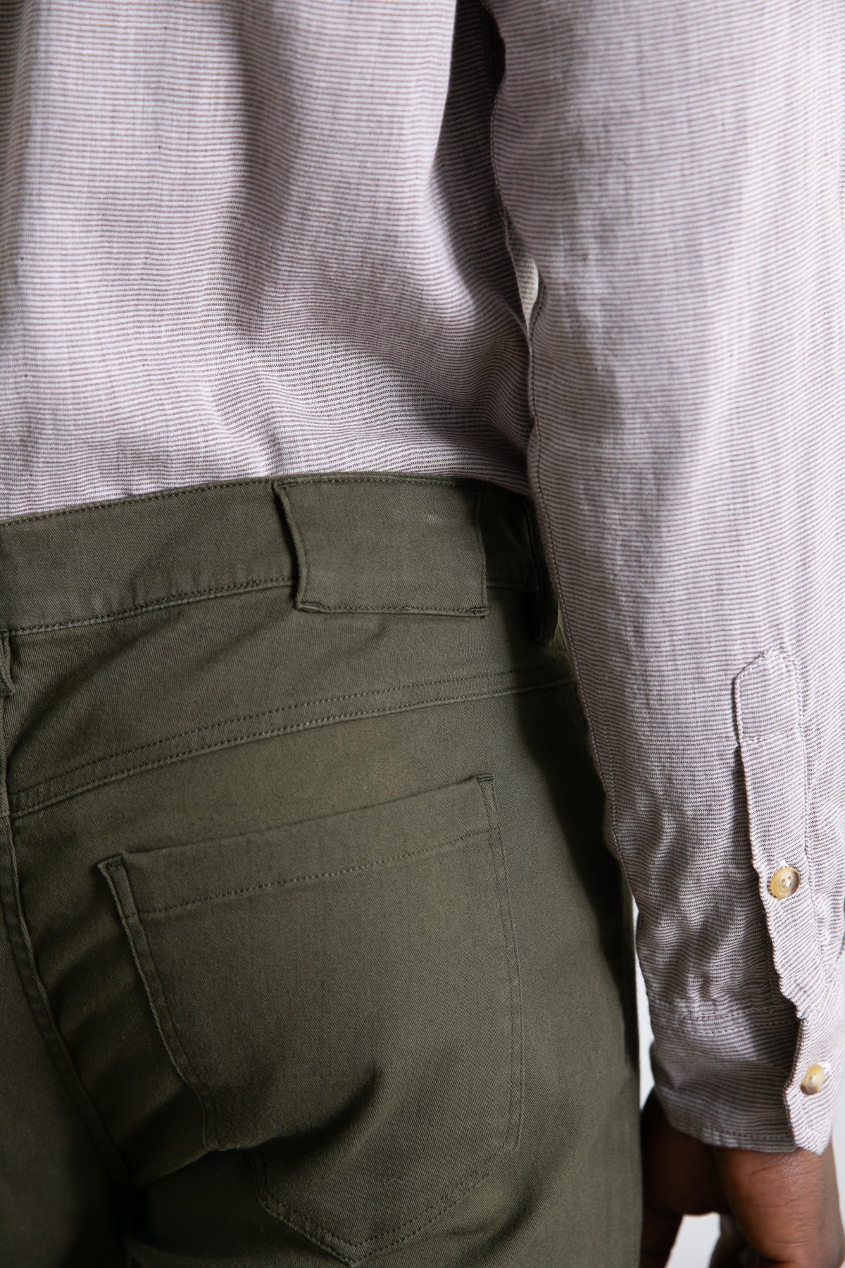 A close-up side view of a person wearing Bridge & Burn's Polk Pant in olive, featuring straight-fit organic cotton and 5-pocket styling, paired with a light long-sleeved shirt with buttoned cuffs. Only the torso and upper legs are shown.