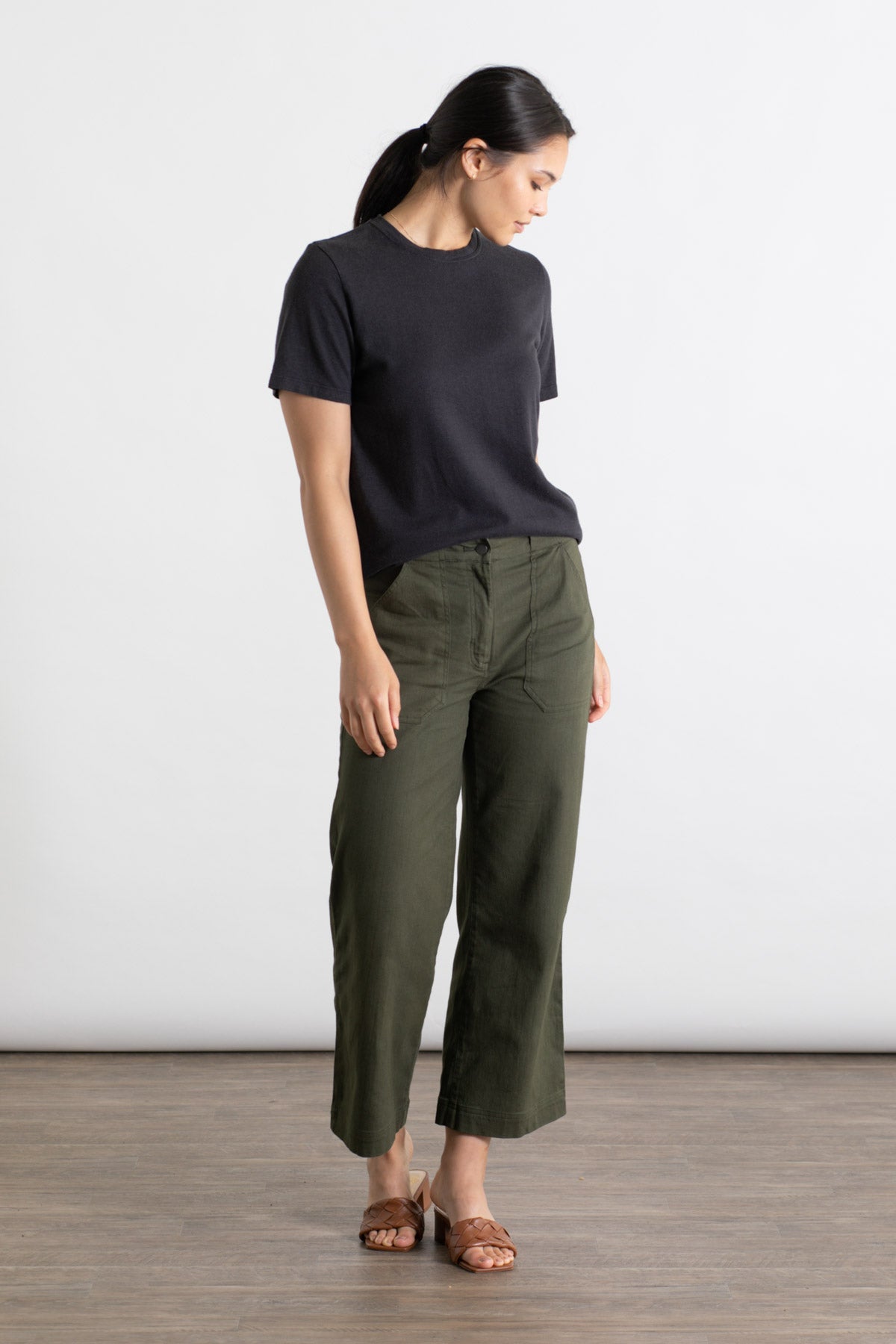 Indoors on a wooden floor, a person with dark hair tied back looks down to their left, wearing the Jungmaven Ojai Tee in black by Jungmaven, olive green wide-leg pants, and brown sandals.