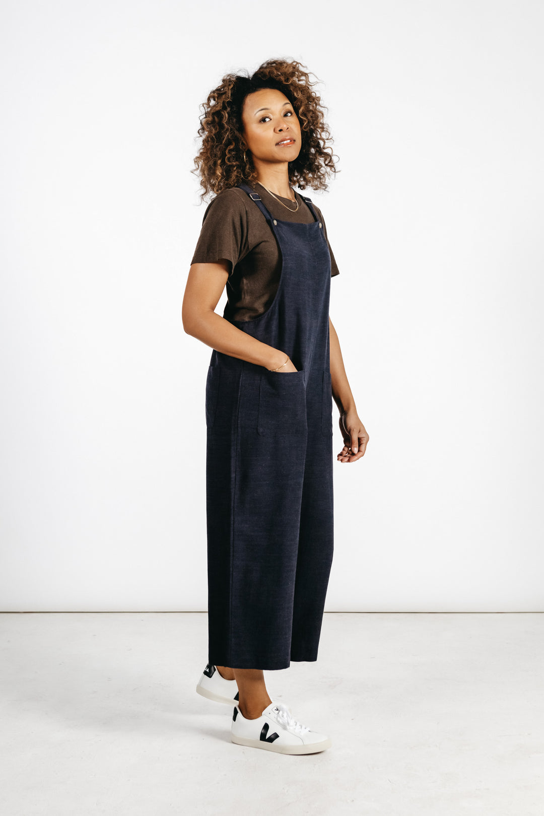 A woman with curly hair stands against a white background, confidently wearing the Bridge & Burn Edie Overall in Navy with a brown t-shirt and classic white sneakers, hands in her pockets.