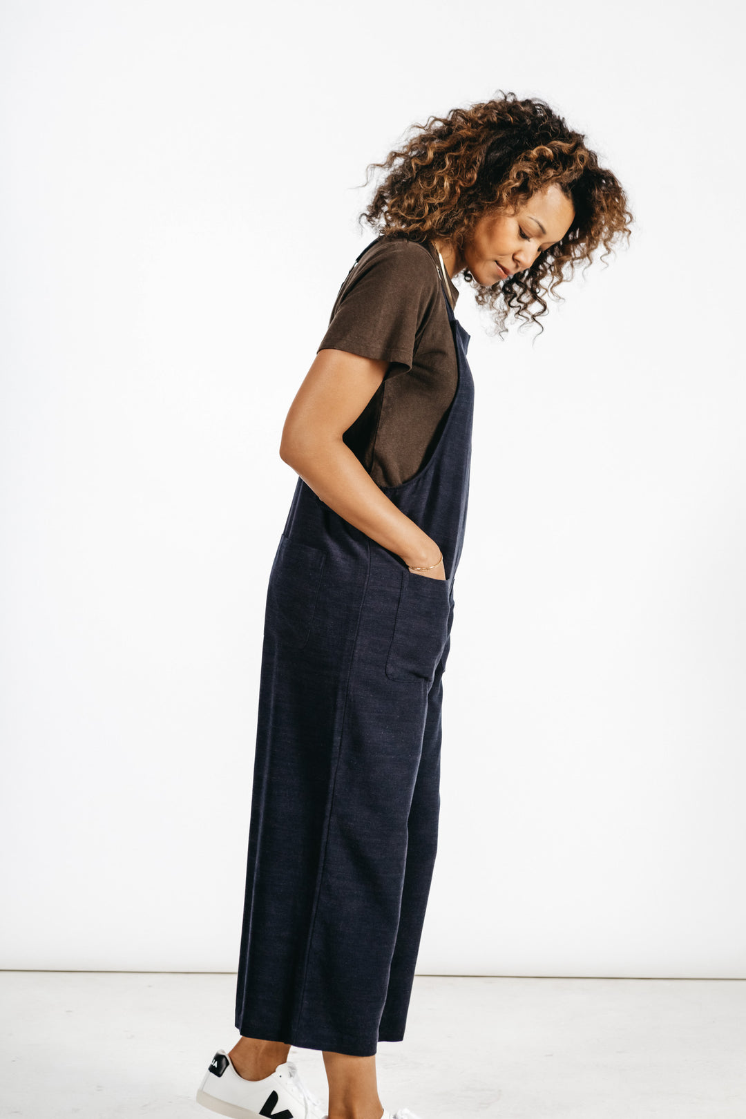 A person with curly hair stands sideways, looking down, wearing the Bridge & Burn Edie Overall in Navy and a brown t-shirt with white sneakers against a plain white background.