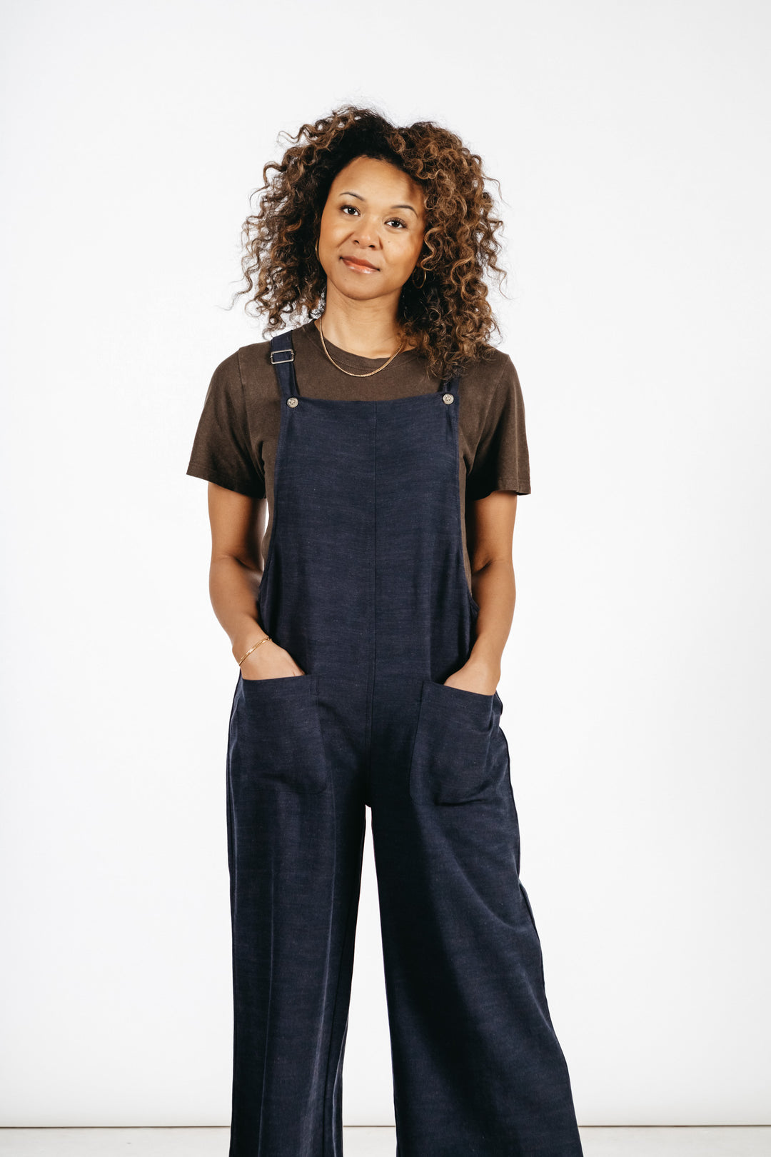 A woman with curly hair wears the Bridge & Burn Edie Overall in Navy with adjustable straps and a dark brown t-shirt, standing hands in pockets and looking slightly to the side against a plain white background.