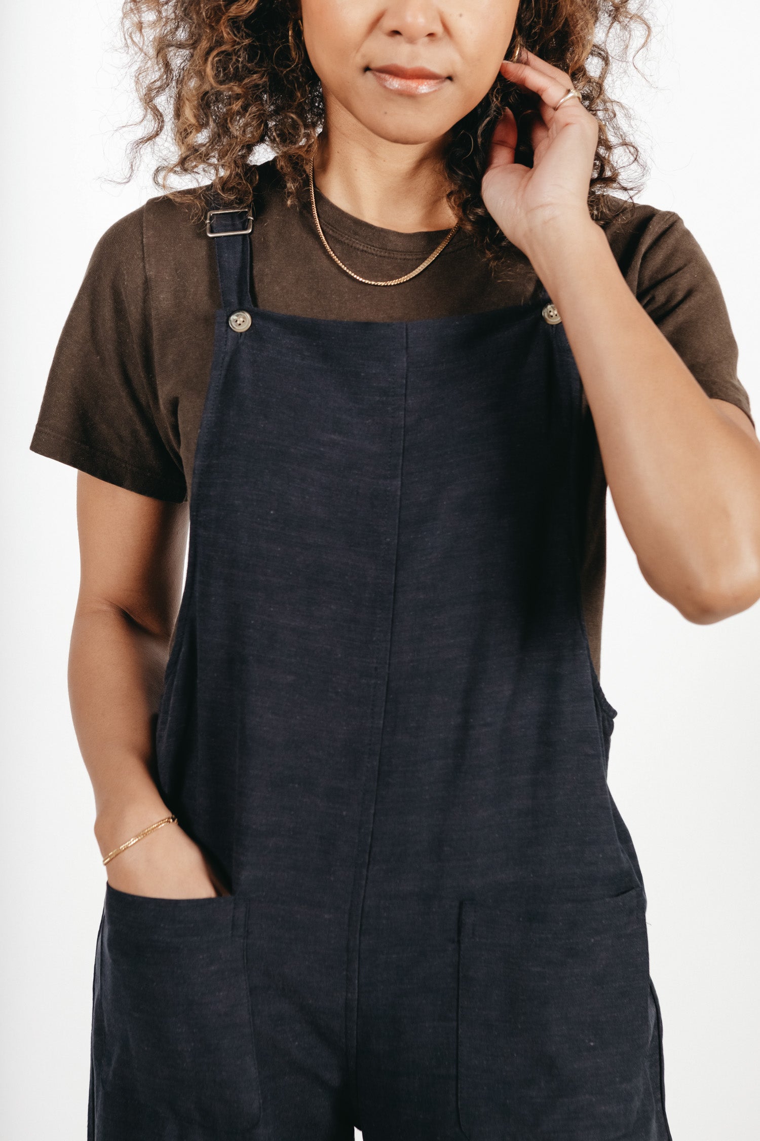 A person with curly hair wears the Bridge & Burn Edie Overall in Navy paired with a dark t-shirt, one hand in a pocket and the other touching their hair, shown from shoulders to mid-thigh against a white background.