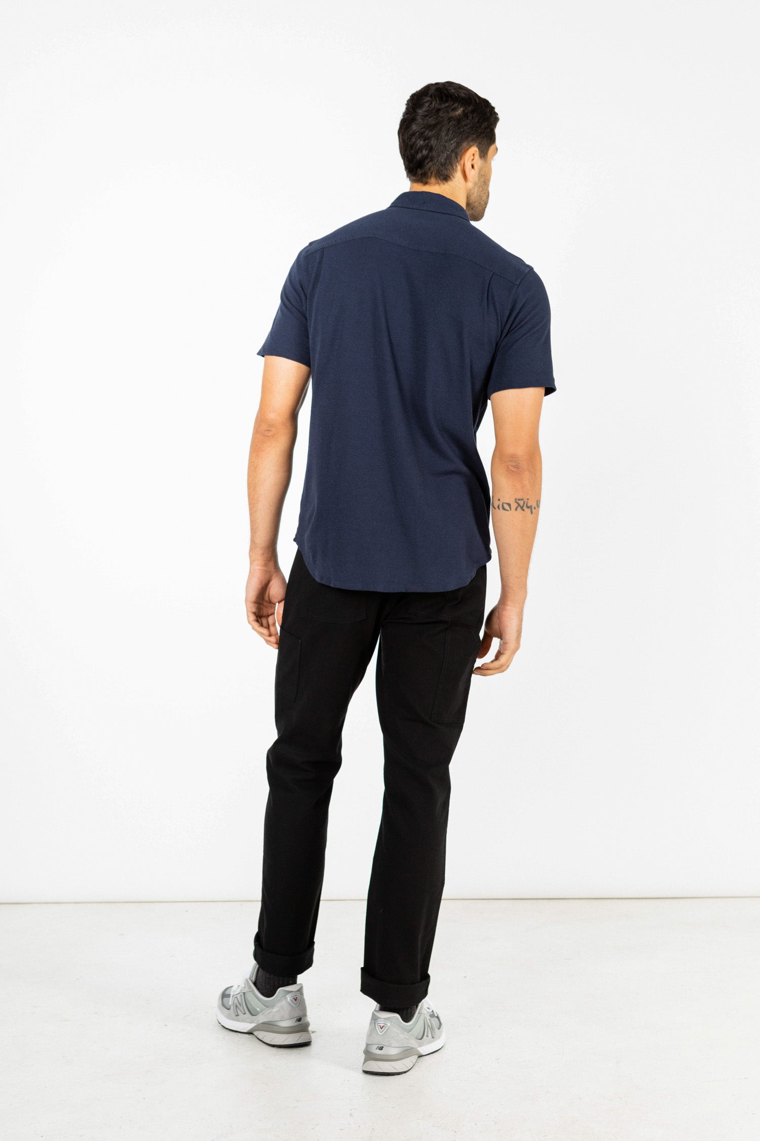 A man faces away from the camera, wearing the Bridge & Burn Gary Knit Pullover in navy jersey and black pants, with gray sneakers, set against a plain white background.