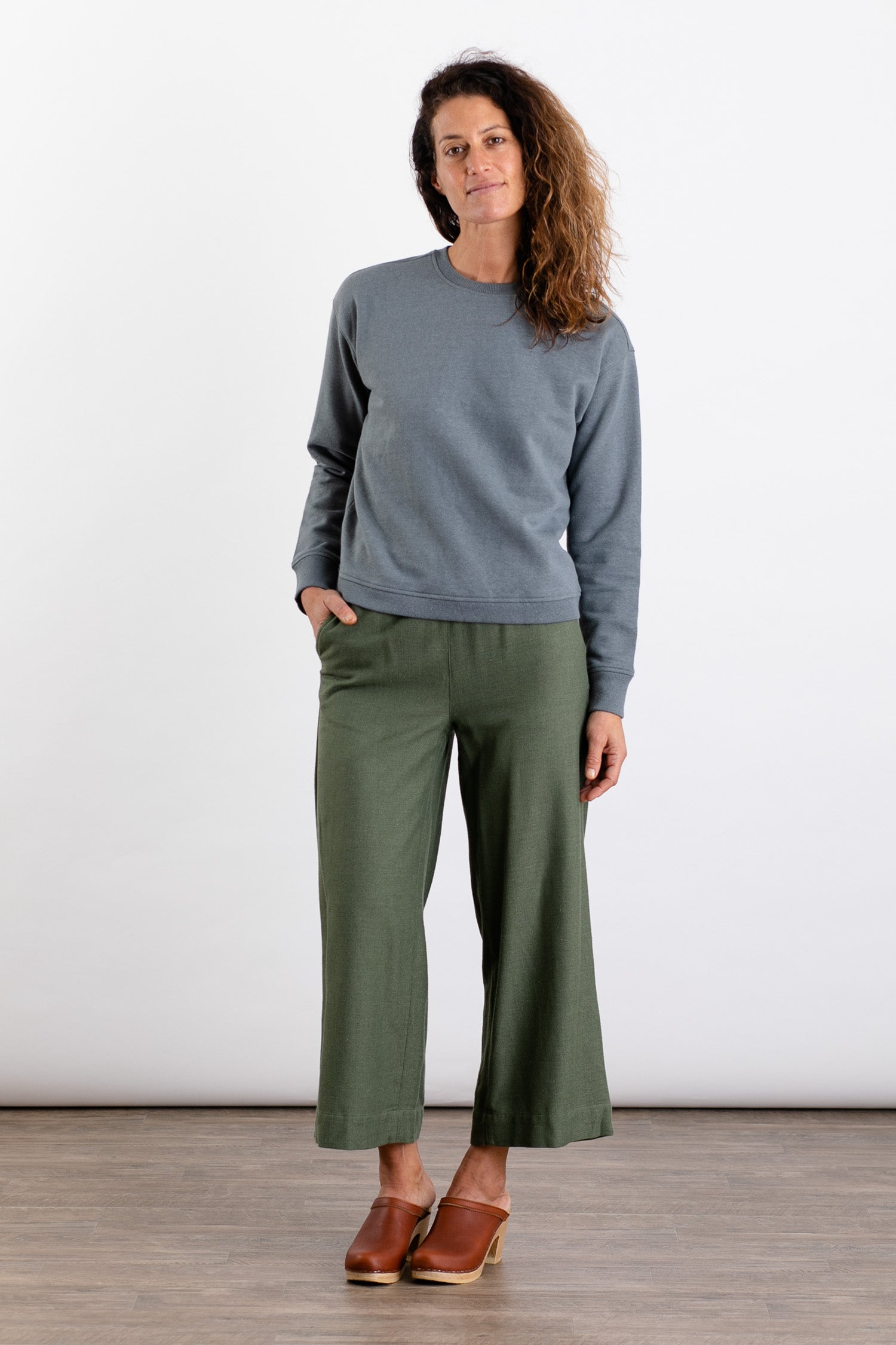A woman with wavy brown hair stands against a white background, wearing the Bridge & Burn Hawthorne Boxy Crew Sweatshirt in Storm Blue, paired with olive green wide-leg pants and brown clogs, one hand in her pocket.