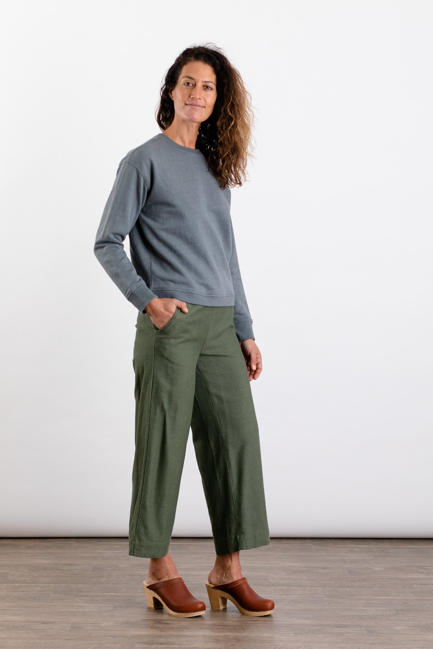 A woman with wavy brown hair smiles in a Bridge & Burn Hawthorne Boxy Crew Sweatshirt in Storm Blue, loose olive pants, and brown clogs. Her left hand is in her pocket as she poses against a plain white background.